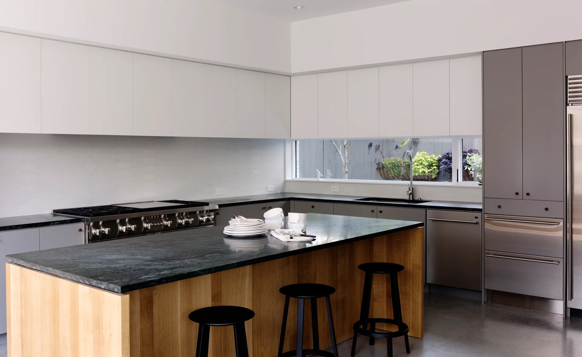 A modern kitchen featuring a wood-toned central island with a dark stone countertop, two-tone flat cabinets, and a horizontal backsplash window.