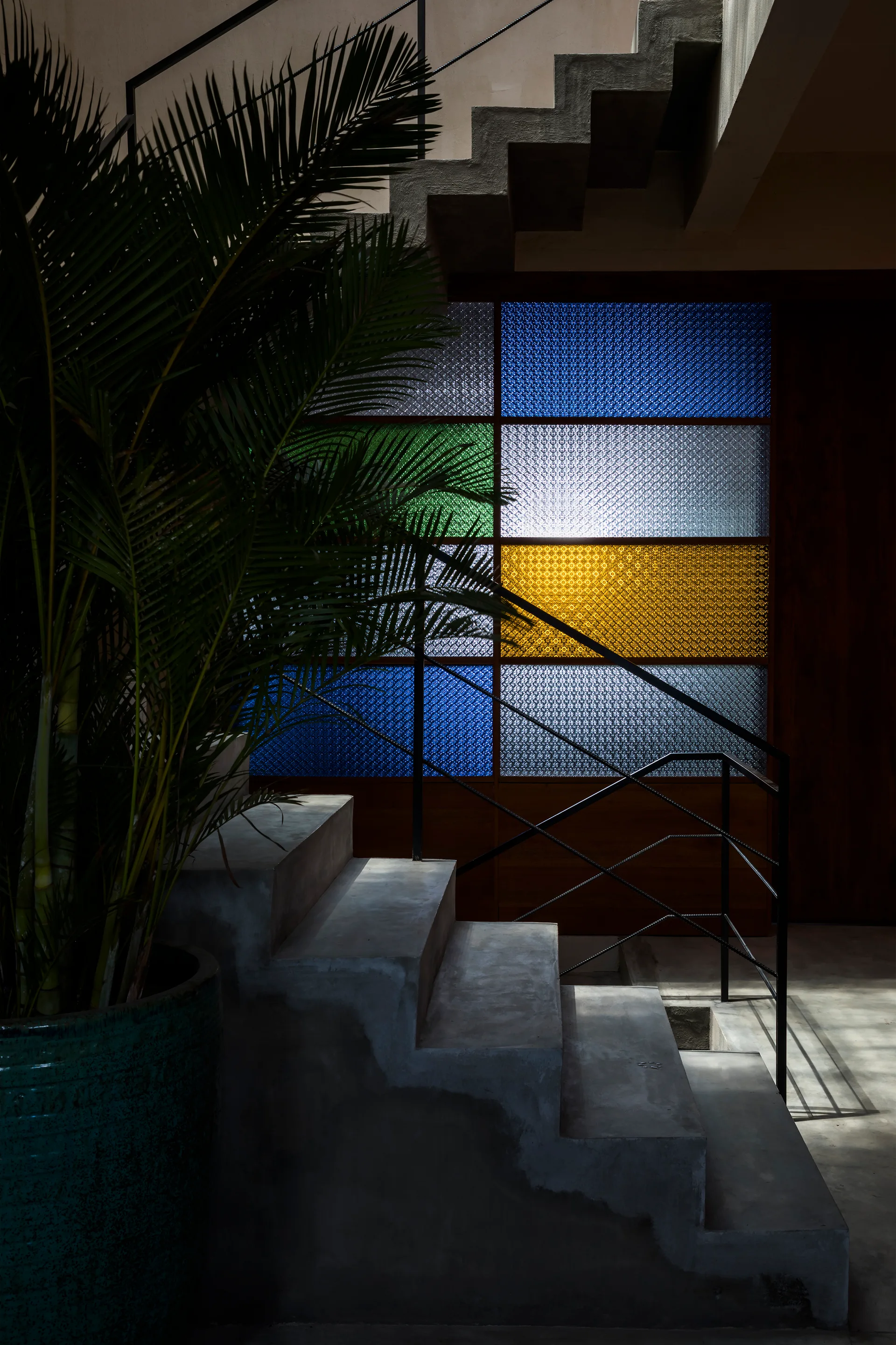 A concrete staircase with black metal railings stands beside a decorative partition wall of blue, green, and yellow textured glass.