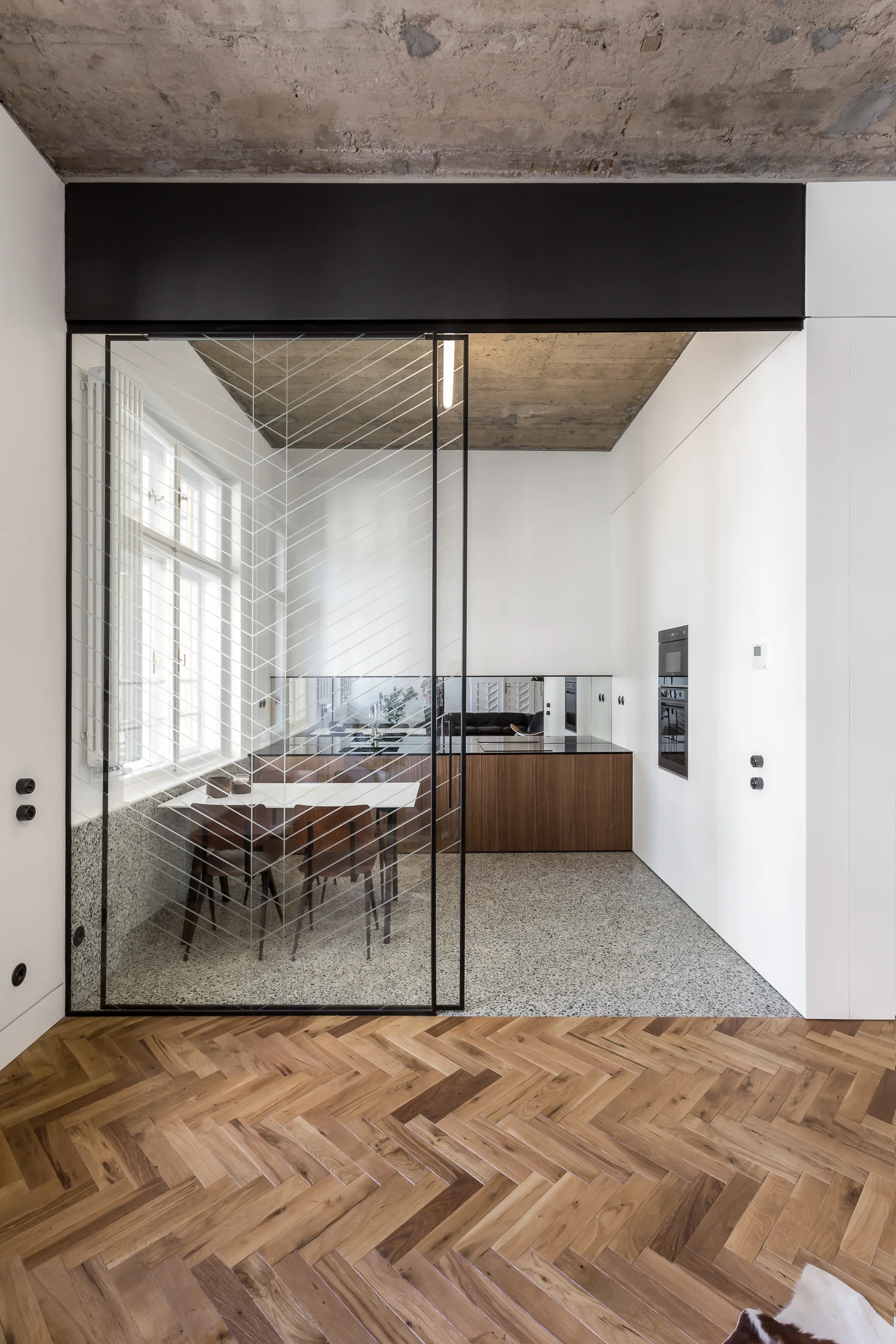 A minimalist kitchen featuring wood cabinets, concrete ceiling, and a glass partition separating herringbone wood and terrazzo flooring.