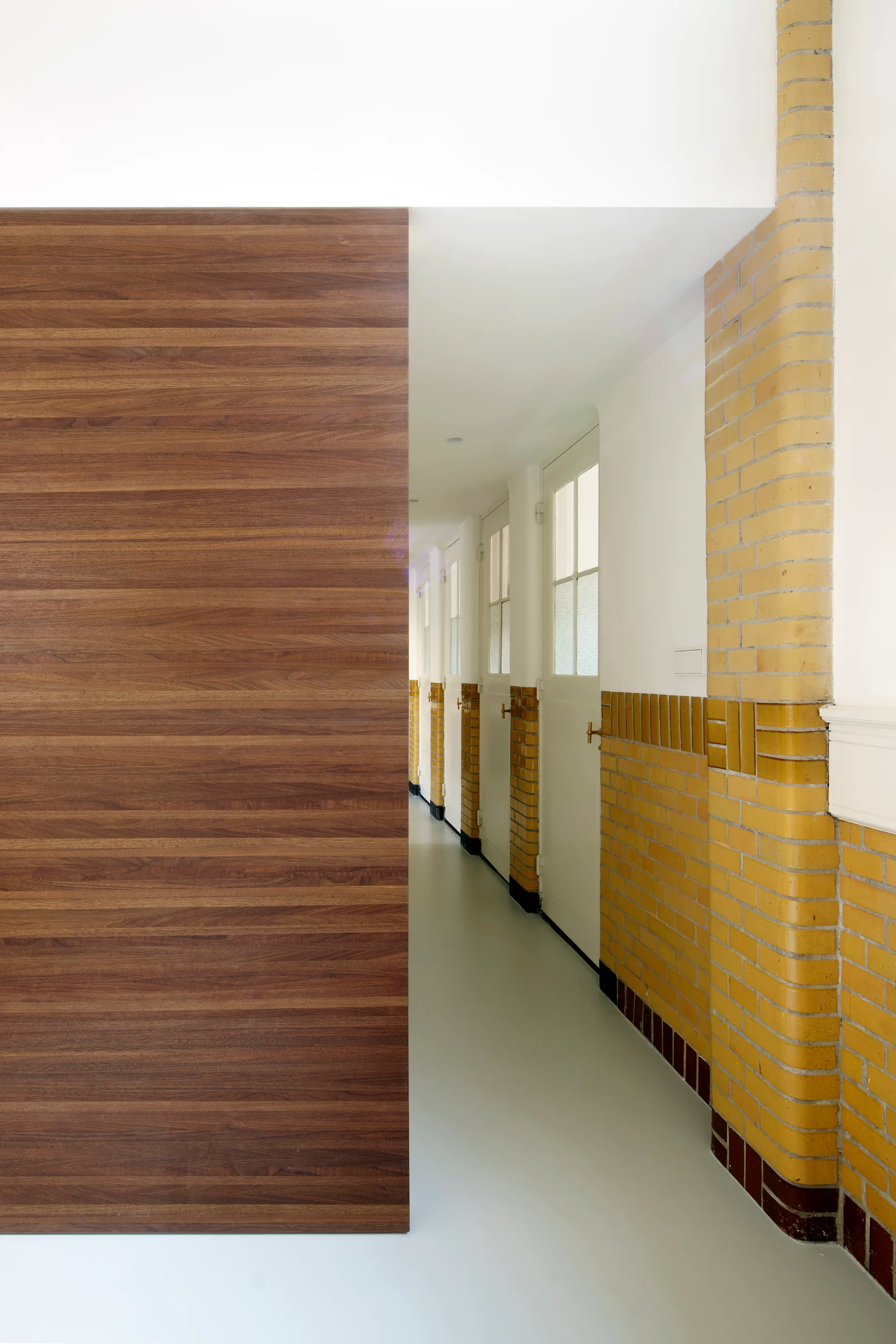 A long hallway featuring a large wood-grain wall panel on the left and glossy yellow brick wainscoting with white paneled doors on the right.