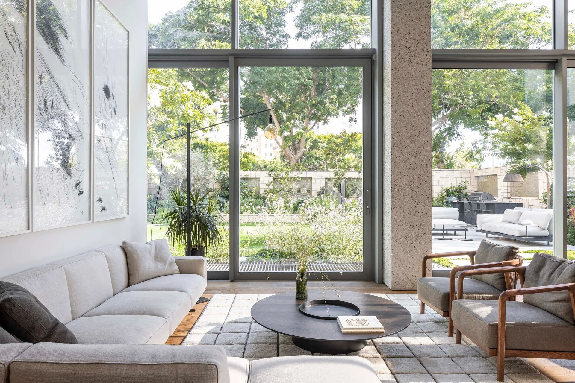 A modern living room featuring a grey sectional sofa, two wooden armchairs, a low round coffee table, and a grid-patterned rug in front of large glass doors.