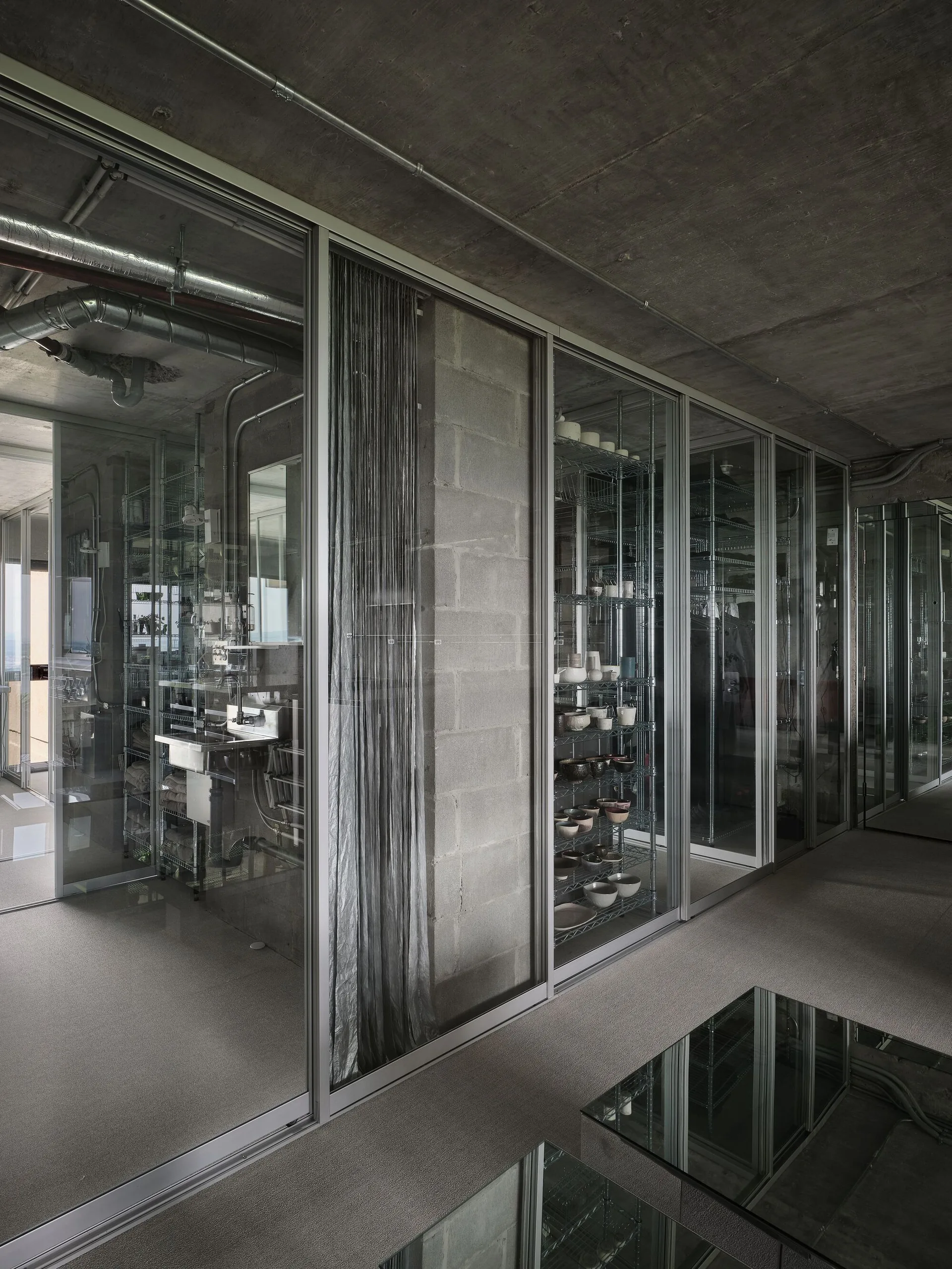 Full-height sliding glass doors partition an industrial room with raw concrete surfaces, metal wire shelving, and an exposed utility sink.