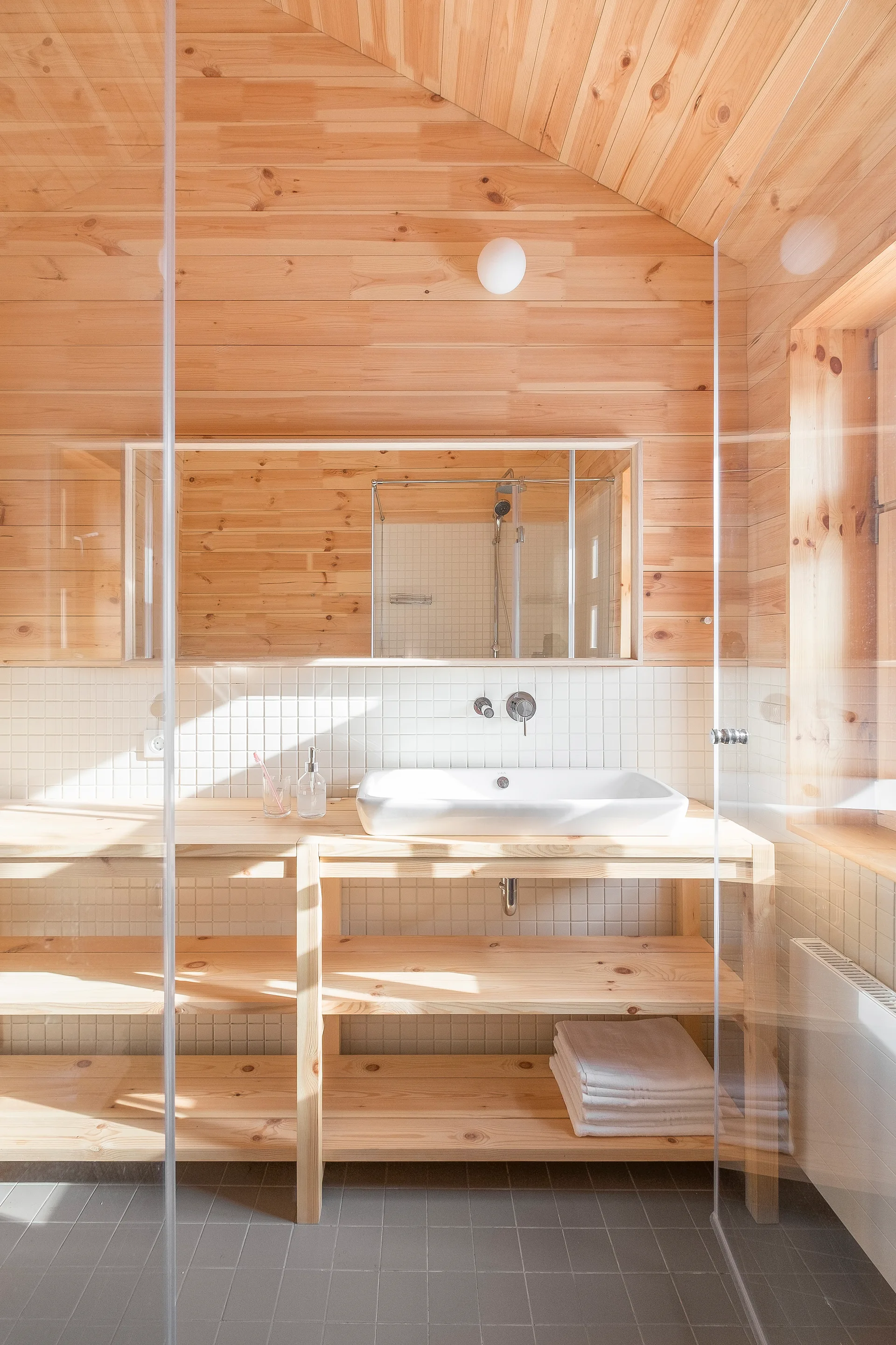 A bright bathroom with natural pine paneling on the walls, a slanted ceiling, a white square-tiled backsplash, and an open wooden vanity.