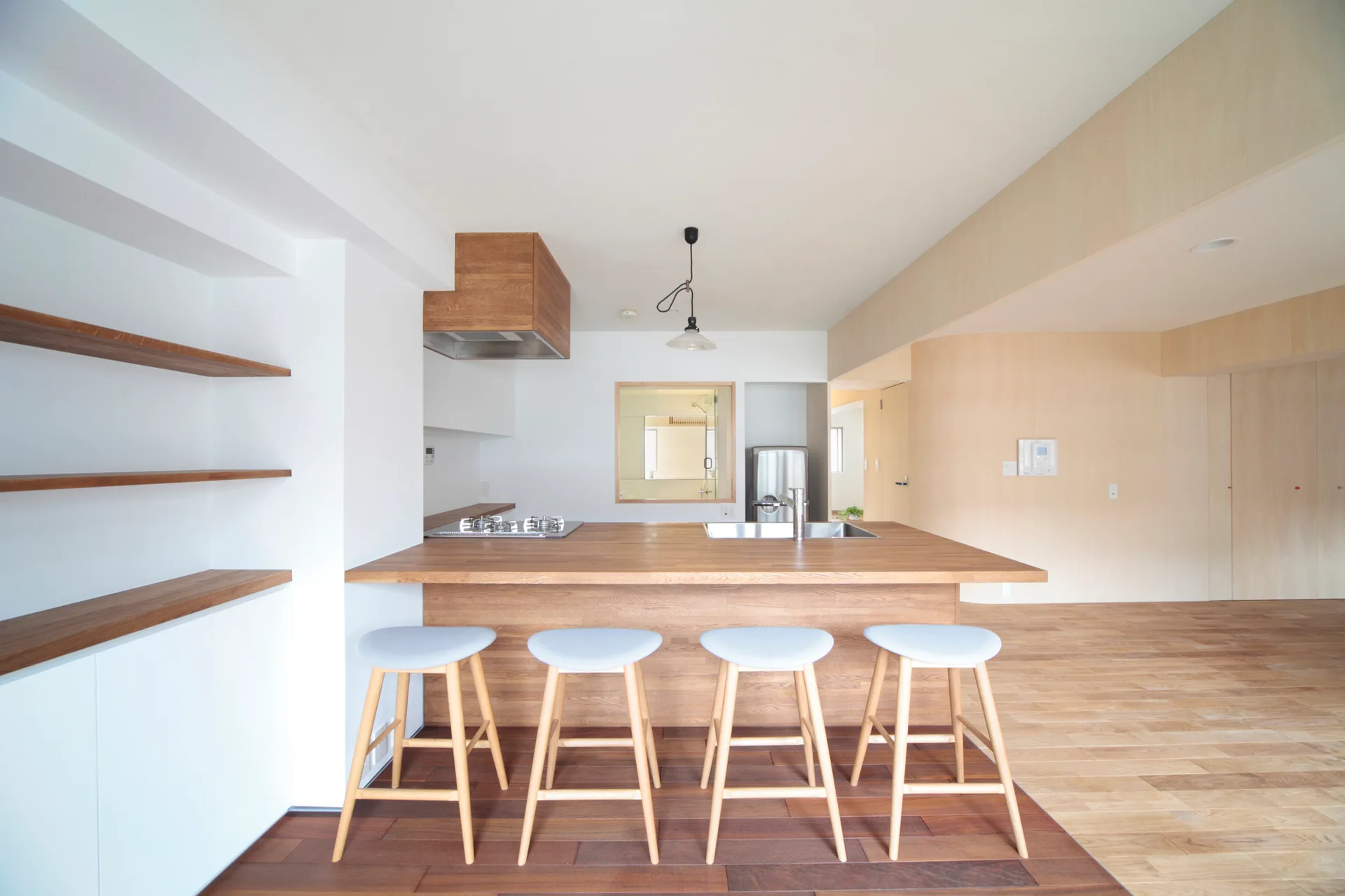 A minimalist kitchen features a wood island with four grey bar stools, open shelving on white walls, and wood-paneled accents.