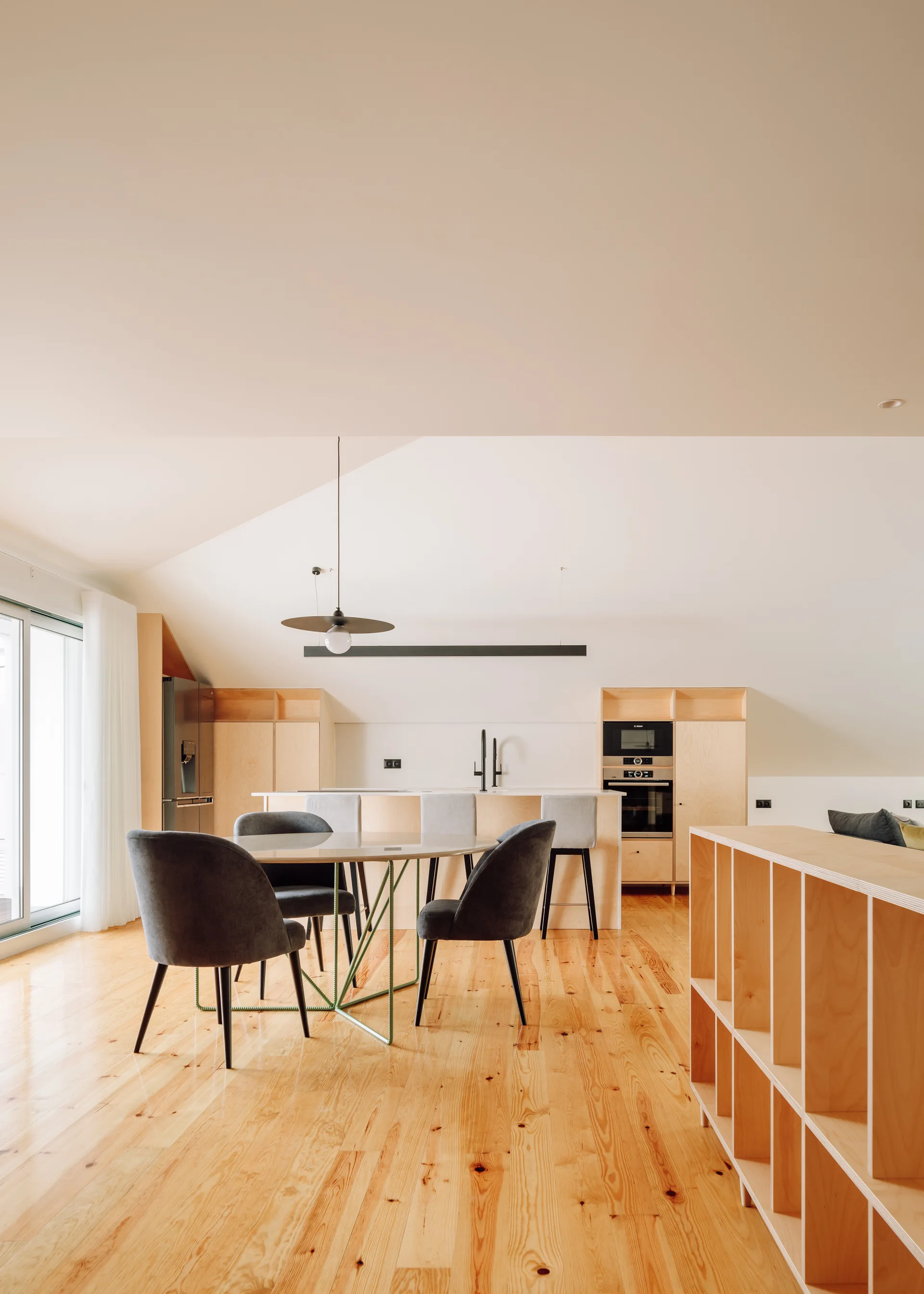 A bright kitchen and dining room featuring light plywood cabinetry, a circular dining table with dark grey chairs, and pine wood flooring.
