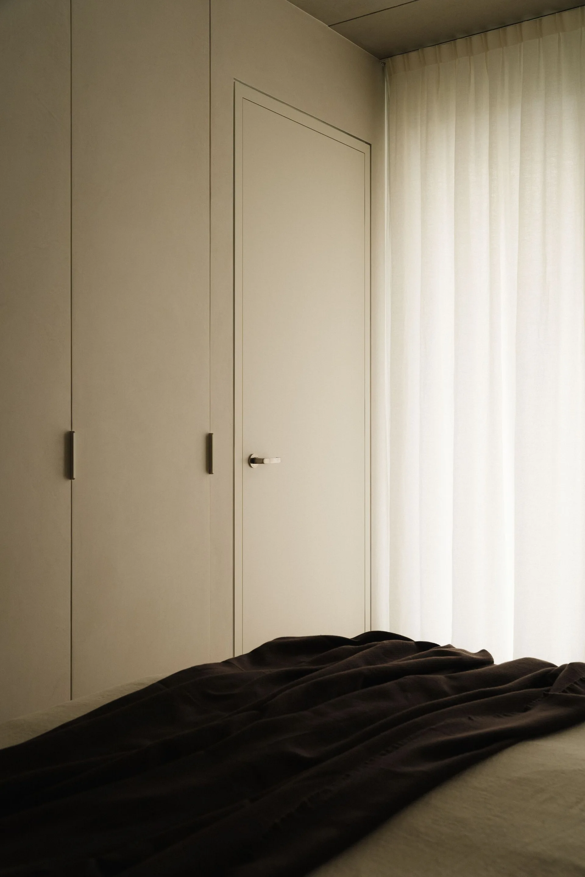A minimalist bedroom featuring seamless warm beige built-in closet doors next to a sheer white curtain with dark bedding in the foreground.