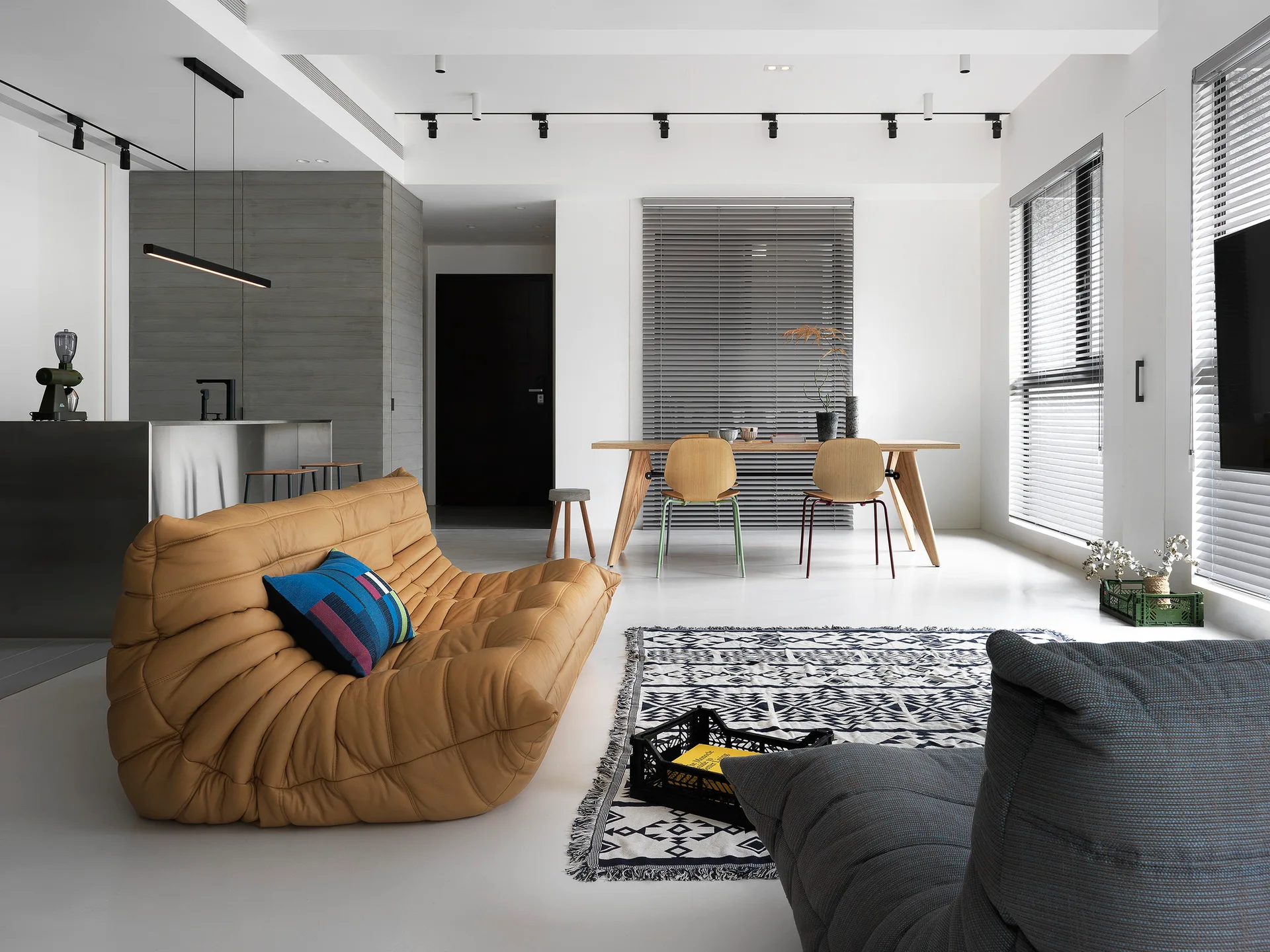 A minimalist open-plan living room features a tufted camel-colored sofa, a geometric patterned rug, and a wooden dining set in the background.