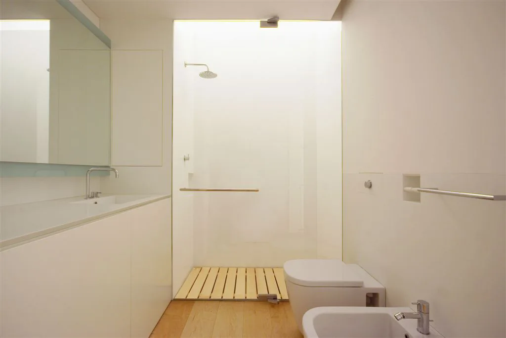 A minimalist white bathroom featuring a seamless vanity, wall-mounted toilet, and a glass-enclosed shower with a slatted light wood floor insert.