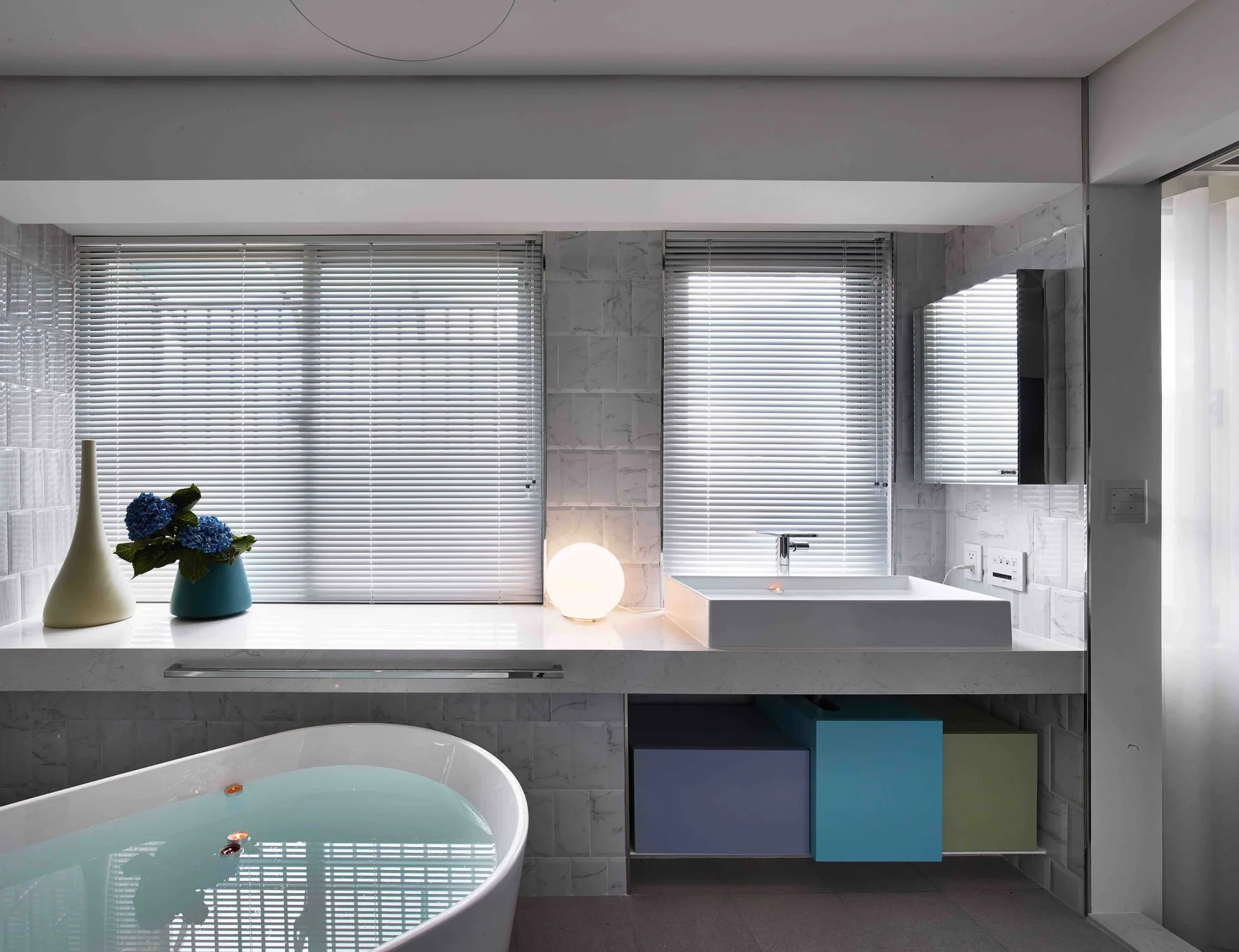A modern bathroom features a white freestanding tub, a marble tiled wall, and a floating vanity with colorful storage cubes underneath.