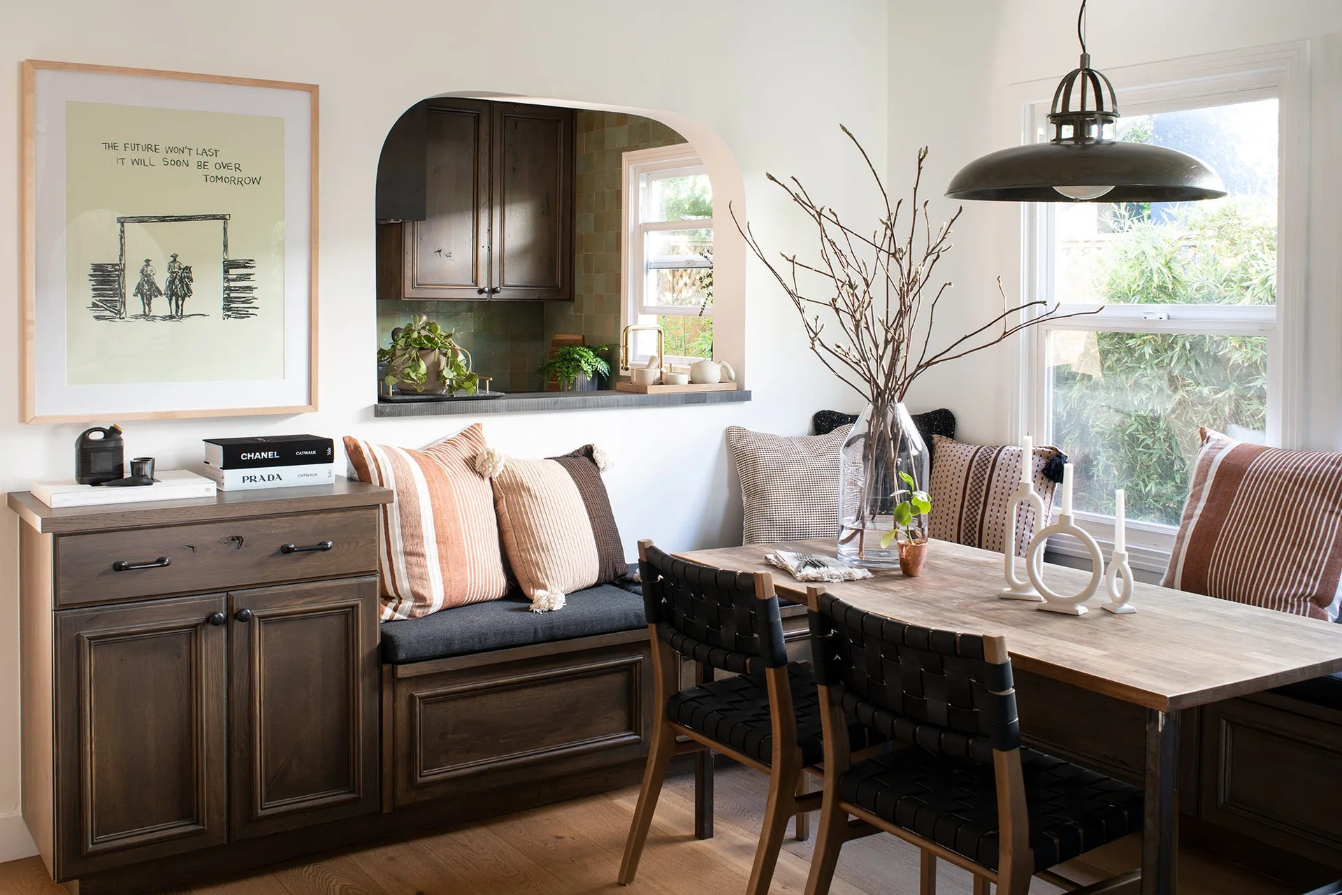 A dining nook features a dark wood built-in bench, a wooden table with woven chairs, and an arched opening looking into the kitchen.