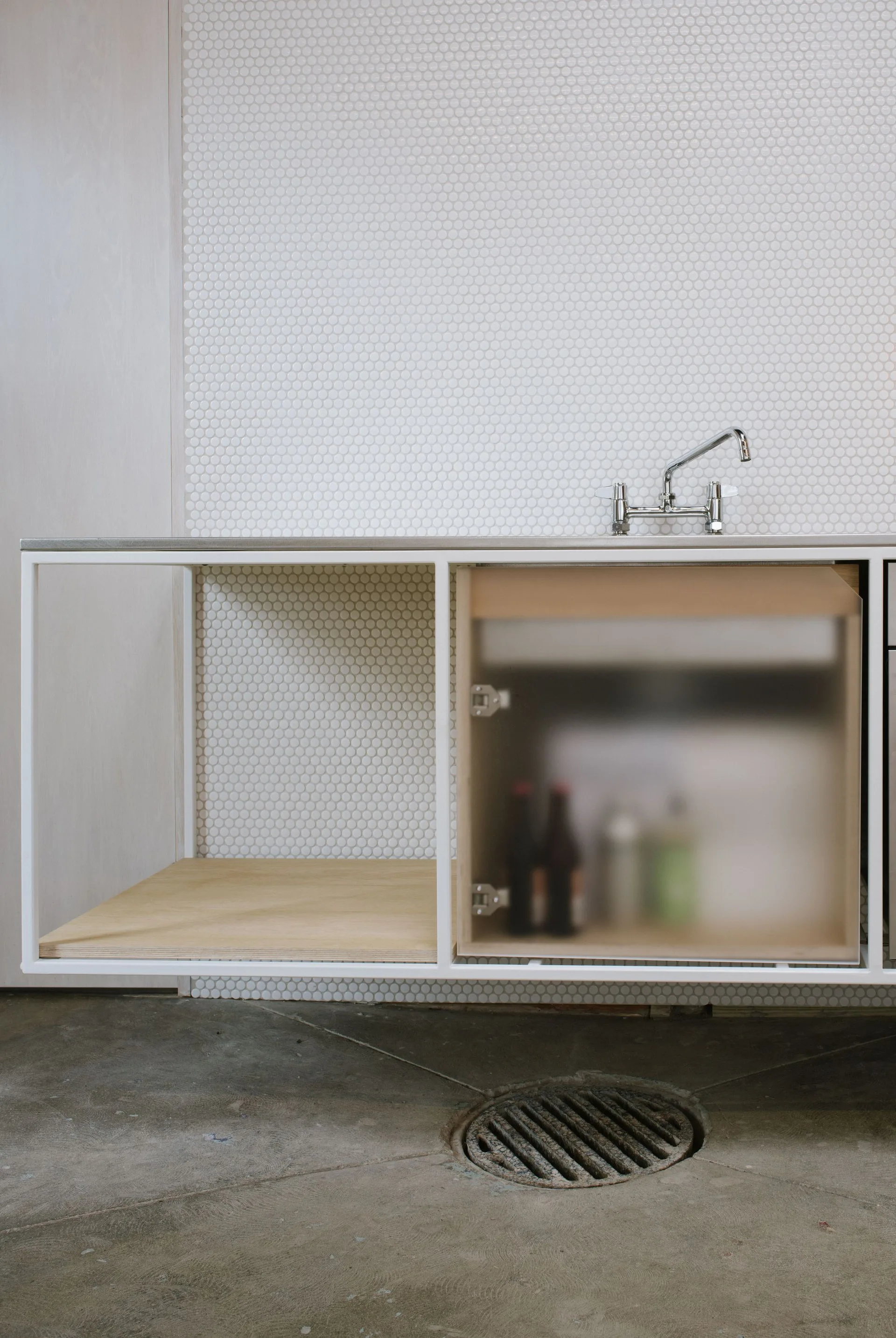 A metal-framed floating vanity features a chrome faucet, frosted glass door, and white penny tile backsplash above a concrete floor.