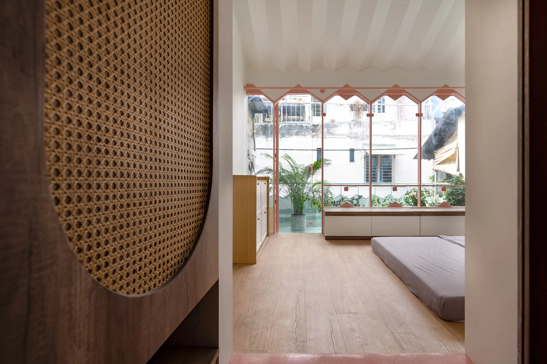 A minimalist bedroom featuring a wooden partition screen with cane webbing, a low platform bed, and large windows with a pink geometric frame.