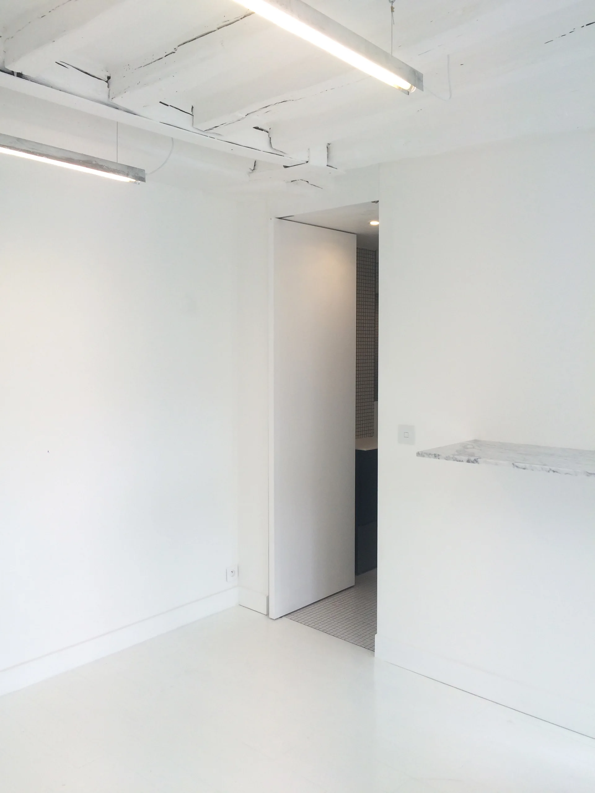 A minimalist white room features exposed painted ceiling beams, a floating marble ledge, and an open doorway leading to a tiled space.