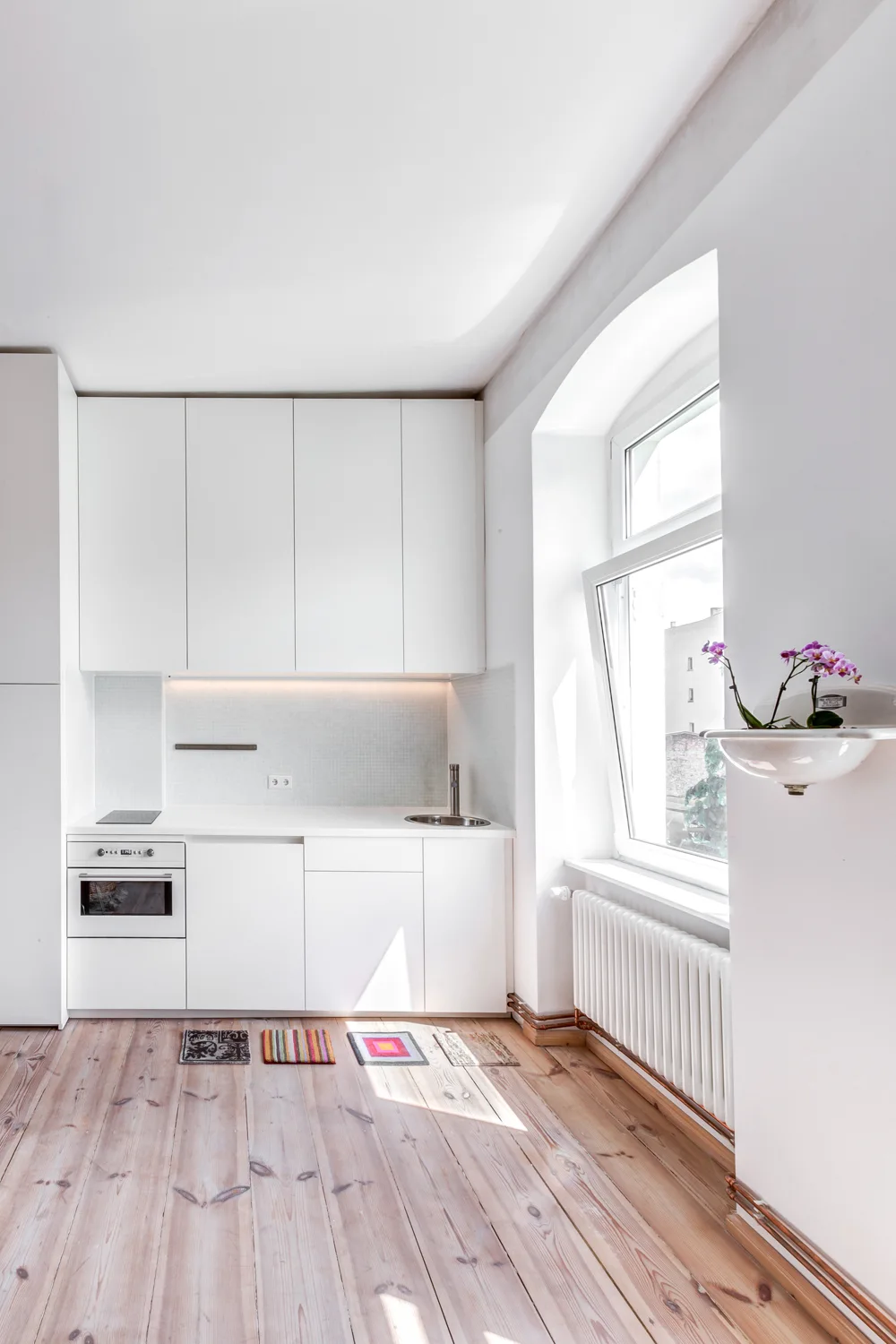 A minimalist kitchen with flat-front white cabinets, a small sink, a tile backsplash, and light wood flooring.