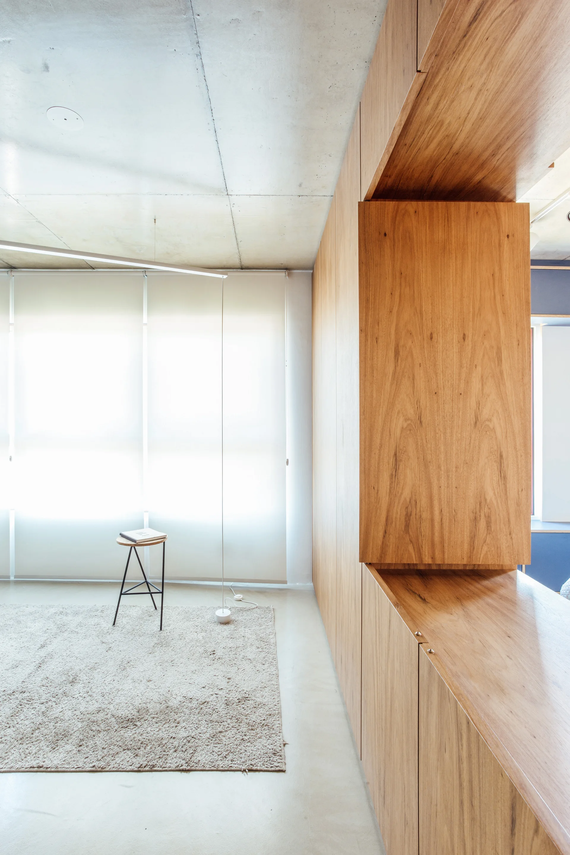 A minimalist room featuring a large custom wood cabinetry unit, a raw concrete ceiling, a textured shag rug, and a small stool.