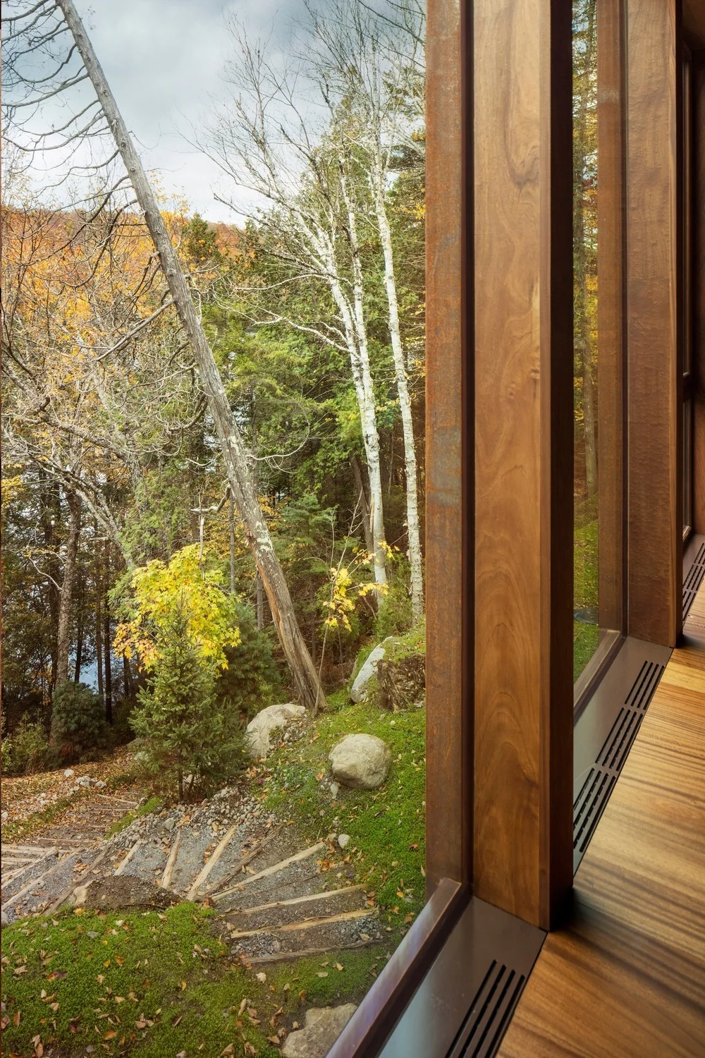 A view through large glass windows framed by thick wood columns looks out onto a green and gold forest with a stepped stone path.