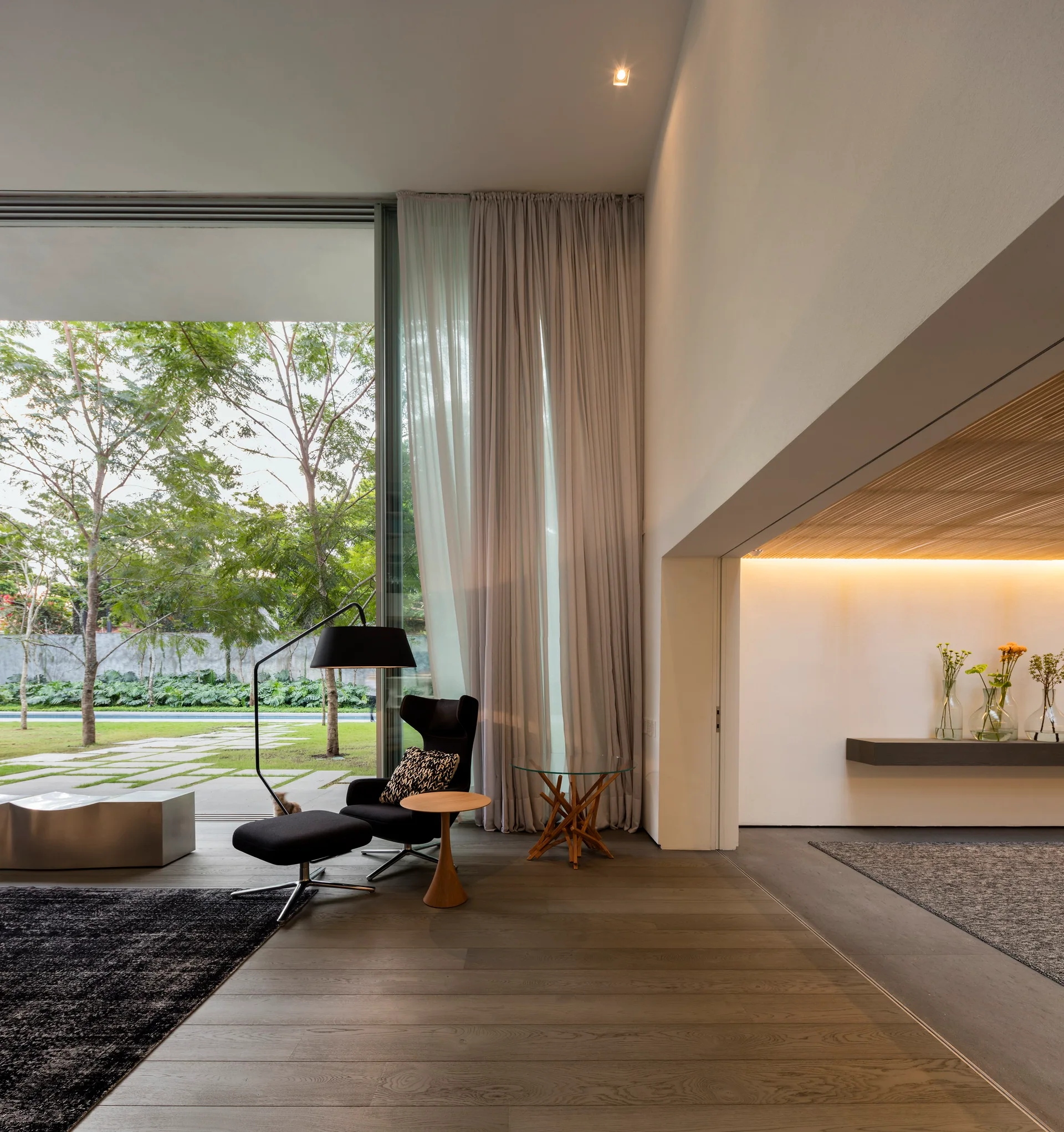 A modern living room features a black lounge chair with an ottoman, towering grey curtains, and large sliding glass doors opening to a lush garden.