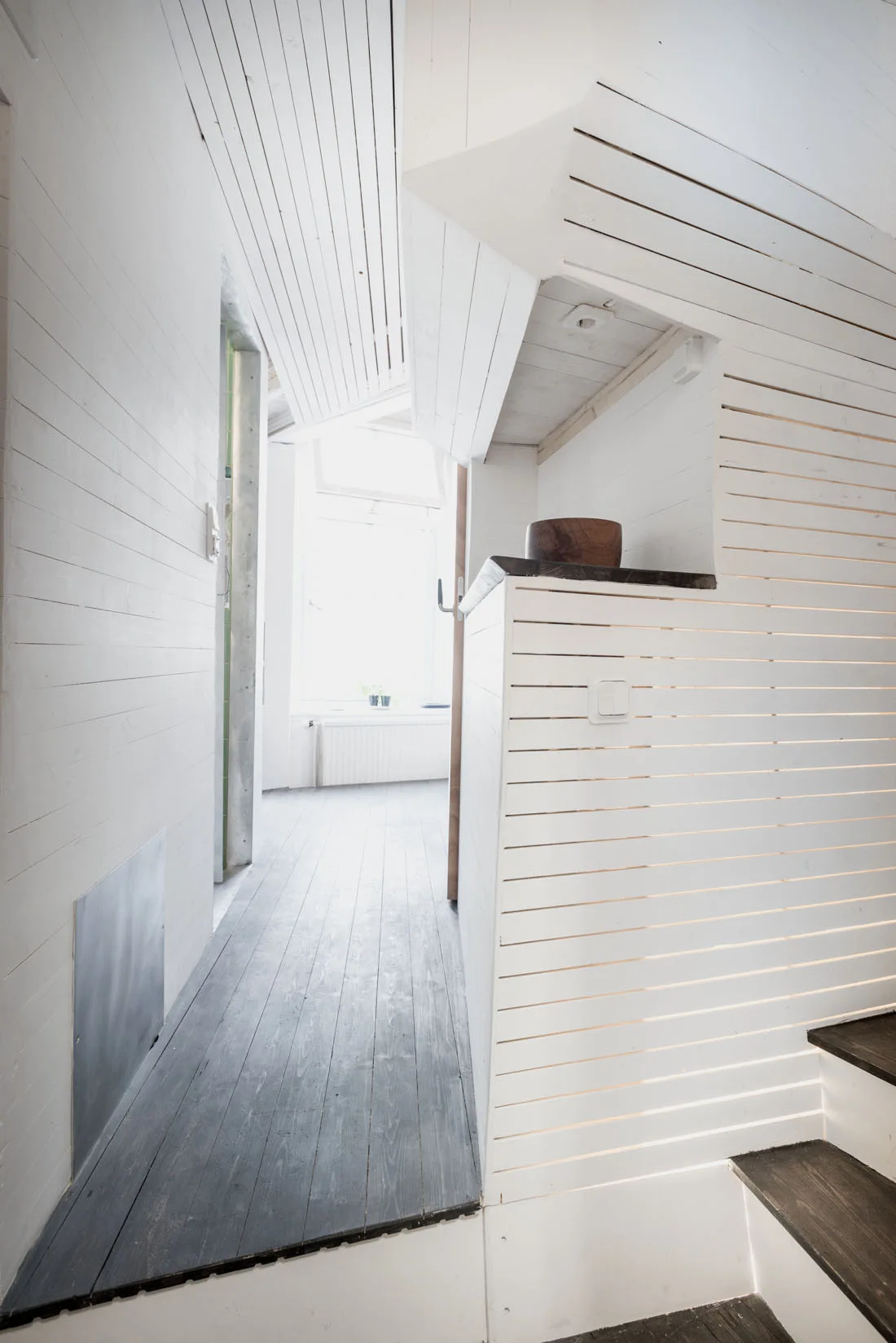 A minimalist white hallway features horizontal slatted wood walls, a dark grey plank floor, and a staircase leading toward a brightly lit window.