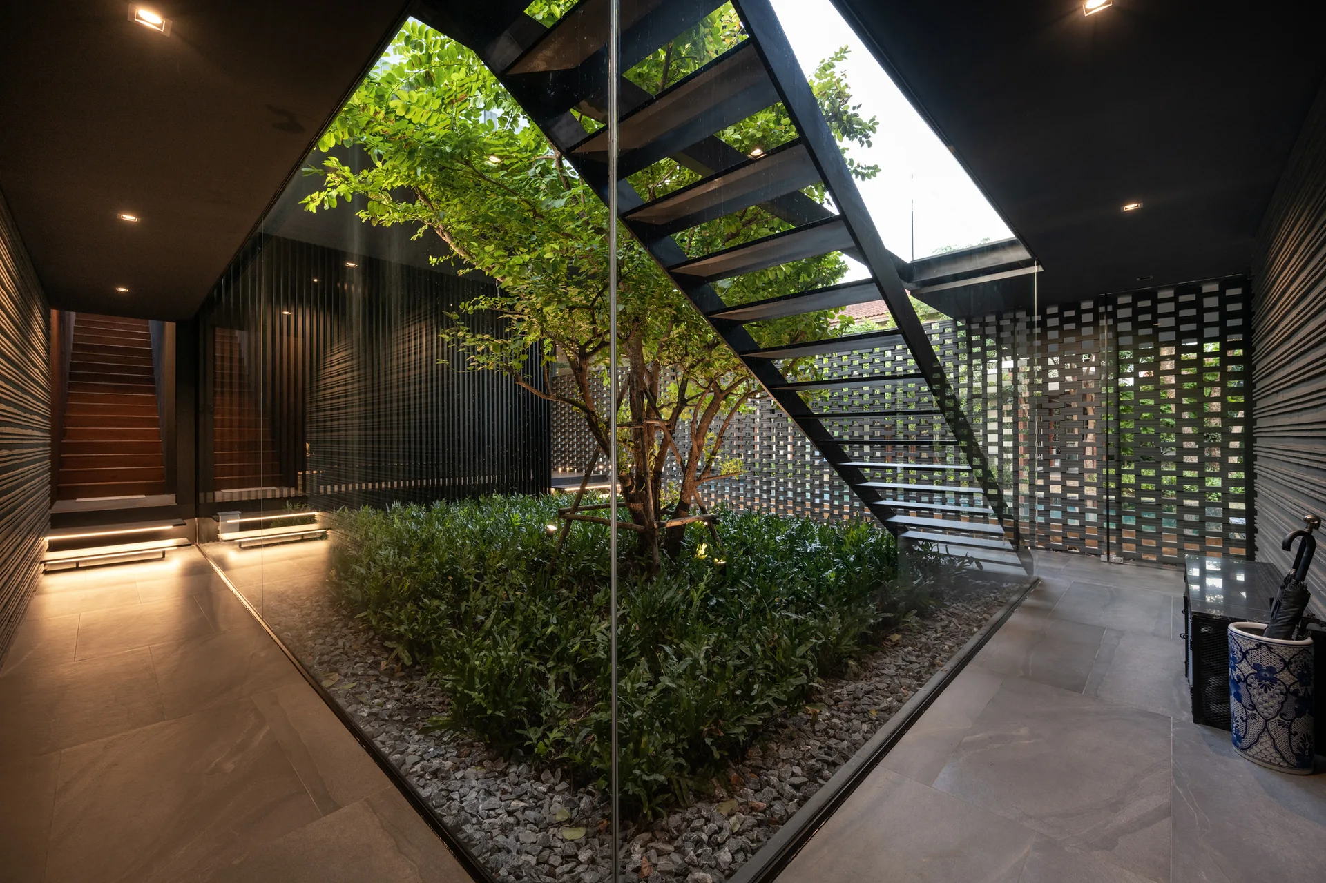 A modern black interior featuring a glass-enclosed central courtyard with a tree, ground cover plants, and a minimalist black metal staircase.