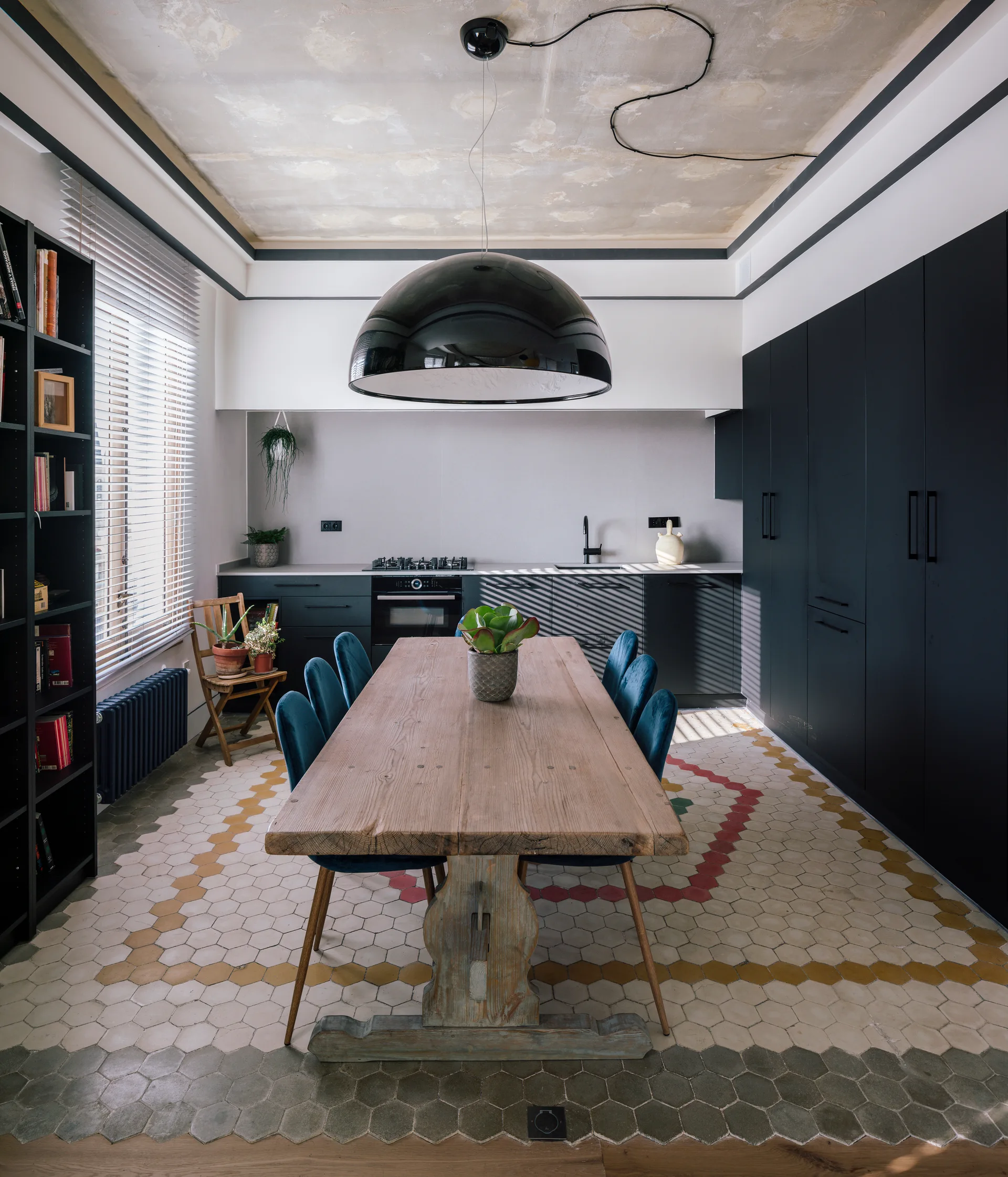 A rustic wood dining table with blue velvet chairs sits on a patterned hexagonal tile floor in front of matte black kitchen cabinets.