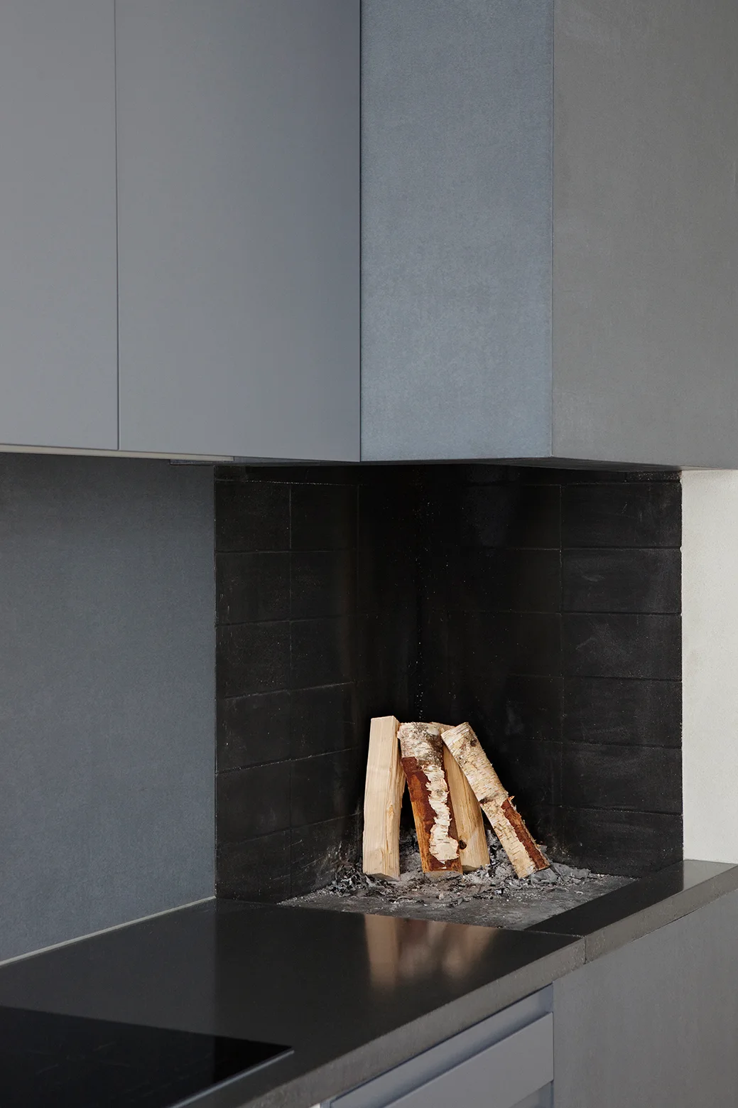 A built-in fireplace featuring black horizontal tiles and birch logs sits beneath matte grey flat-panel kitchen cabinetry.