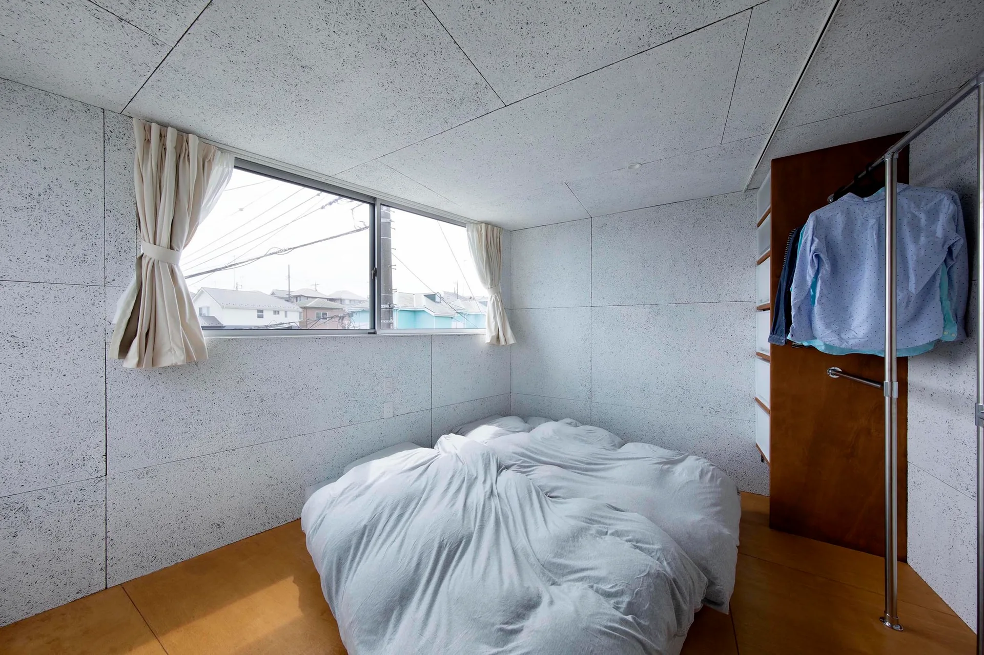 A minimalist bedroom features a low bed with grey linens, textured speckled wall panels, and an open wood wardrobe.