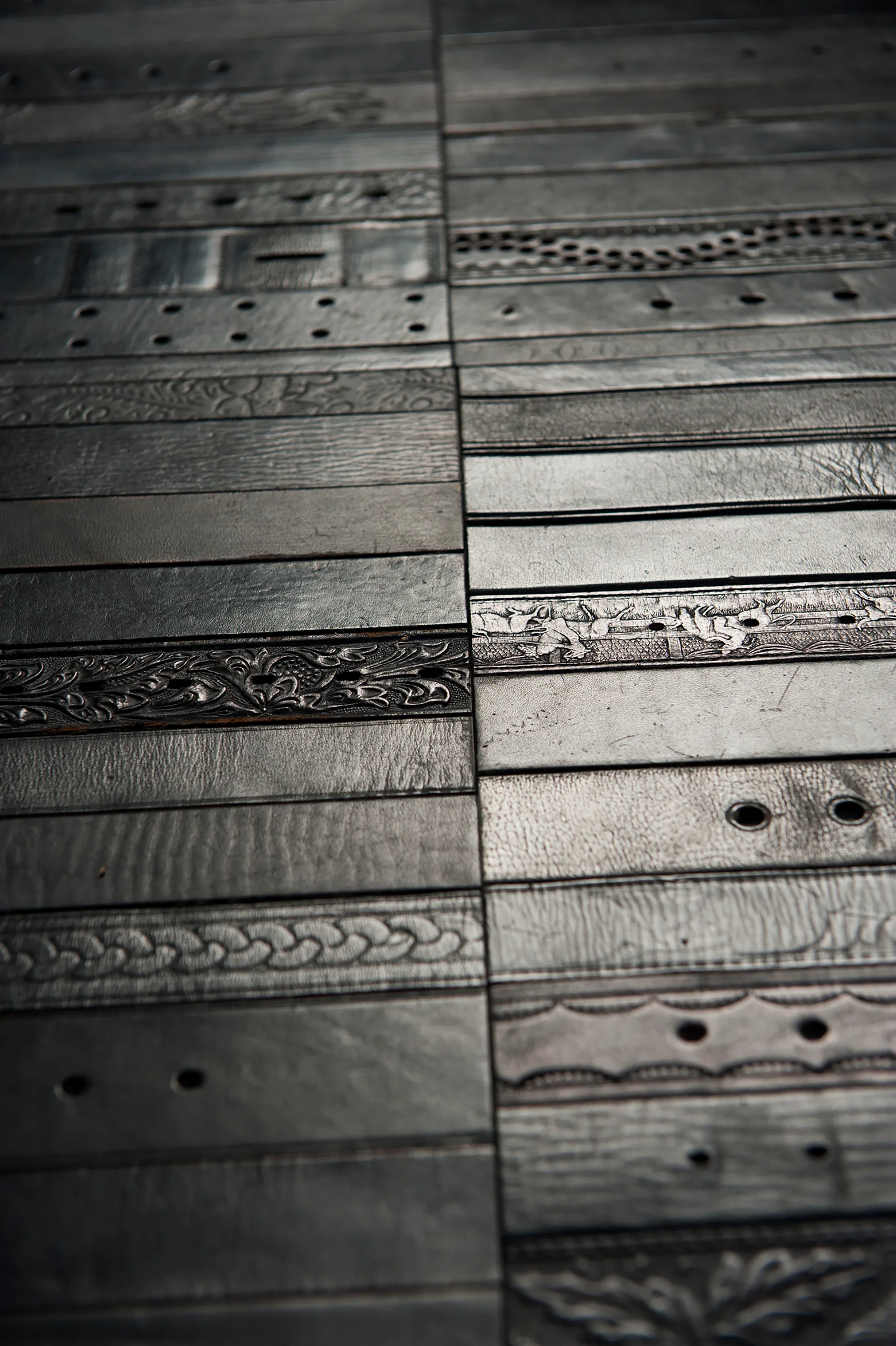 A close-up view of dark brown leather belts with varied embossed floral and geometric patterns laid side by side in parallel rows.