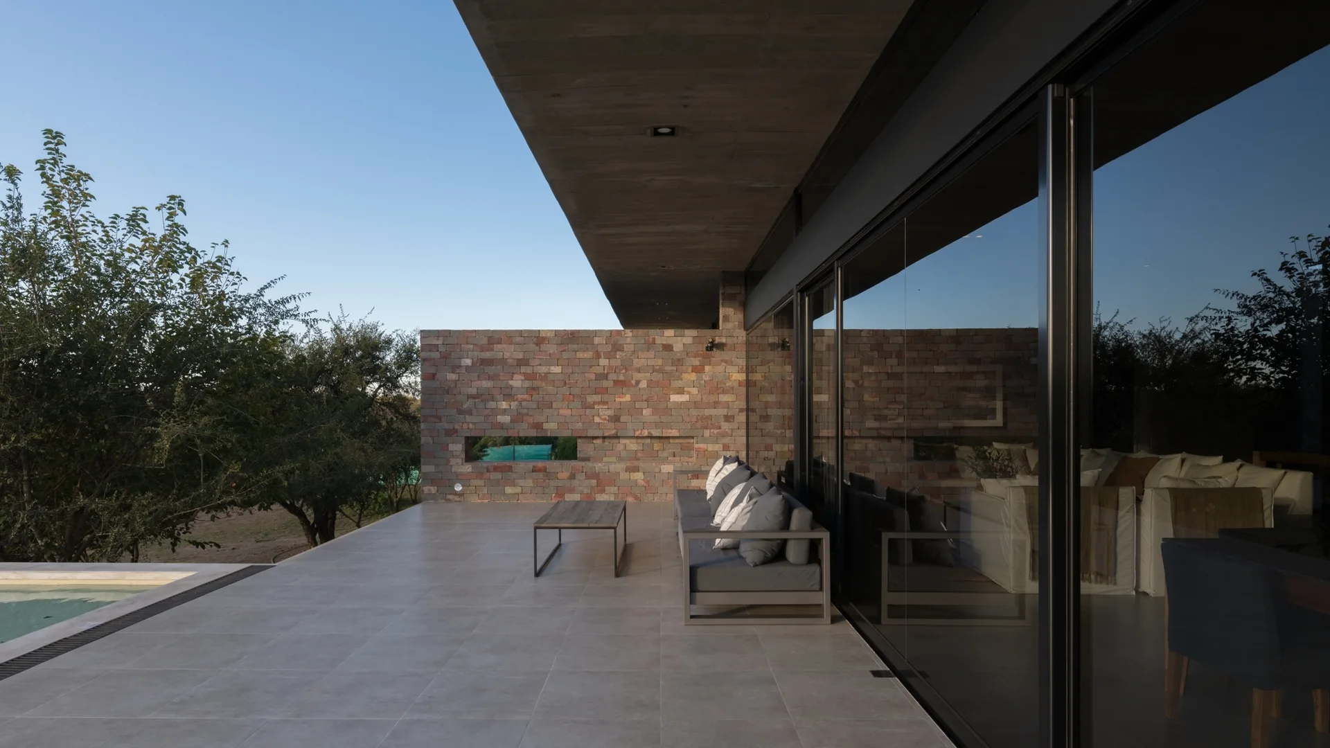 A modern patio with a grey sofa and coffee table sits under a concrete overhang next to a multicolored stone wall and pool.