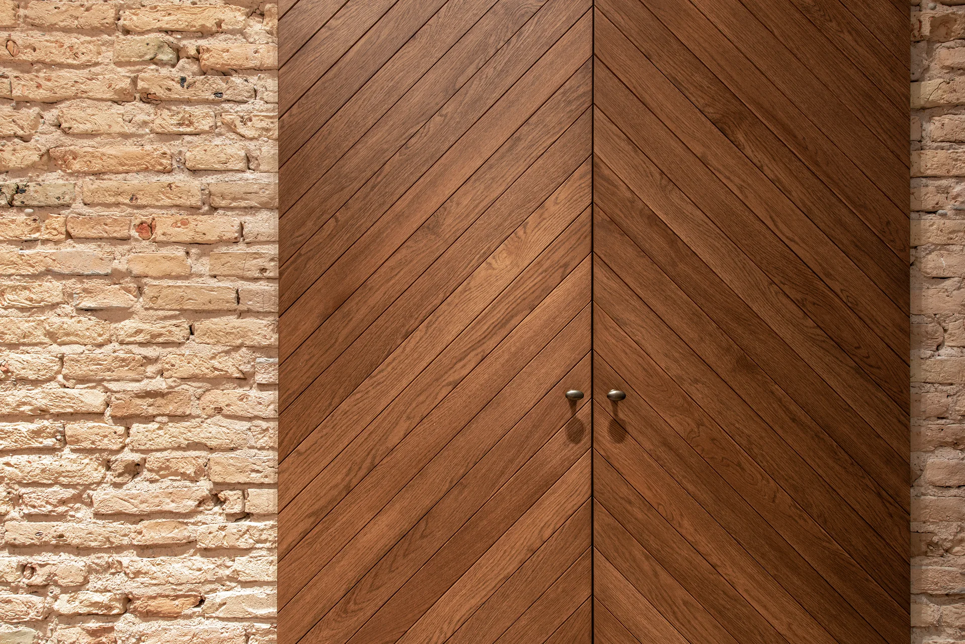 Dark brown wood double doors with a diagonal chevron pattern set against a rough beige brick wall.