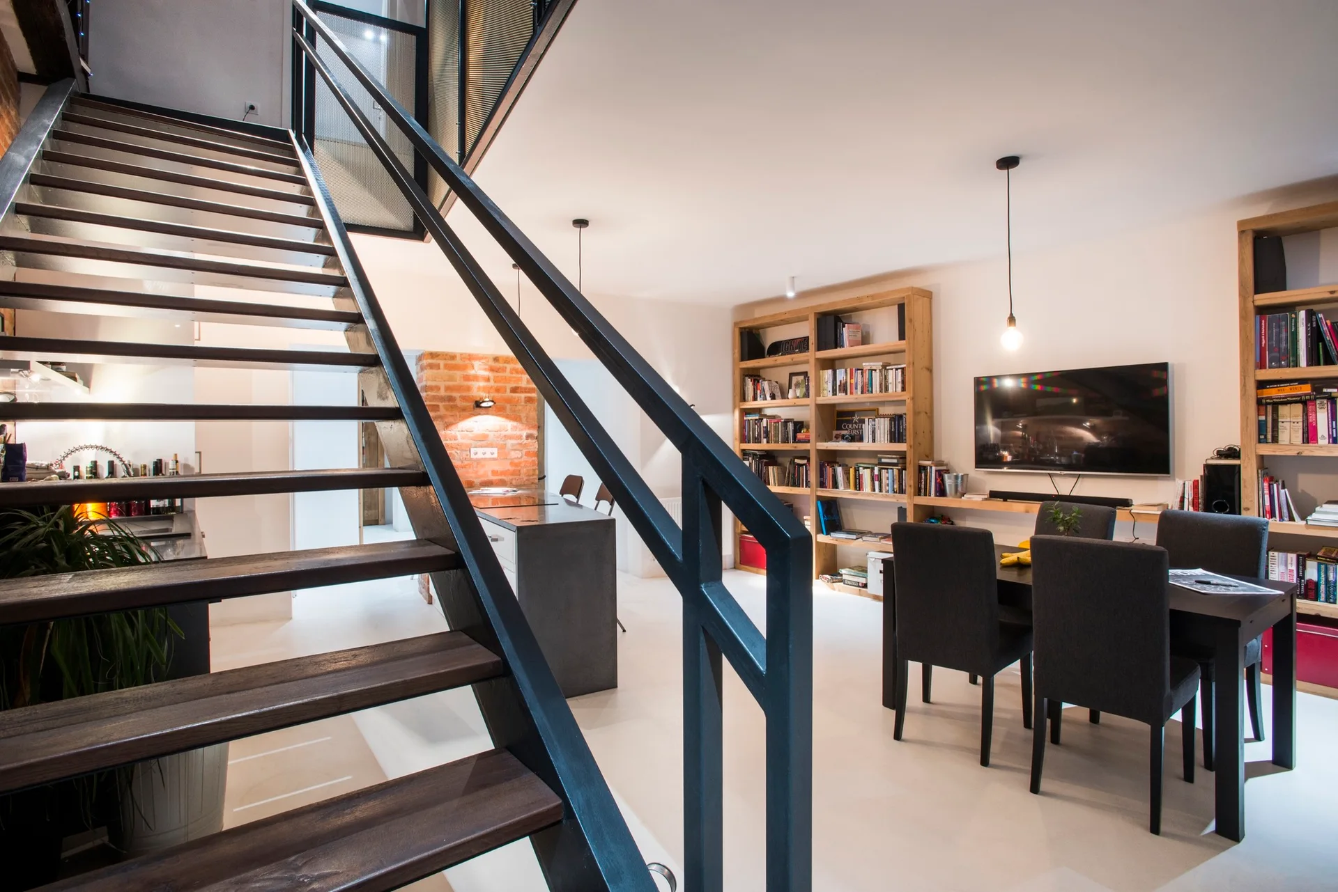 An industrial black metal staircase with wood treads stands in an open room with a dark dining table and light wood bookshelves.