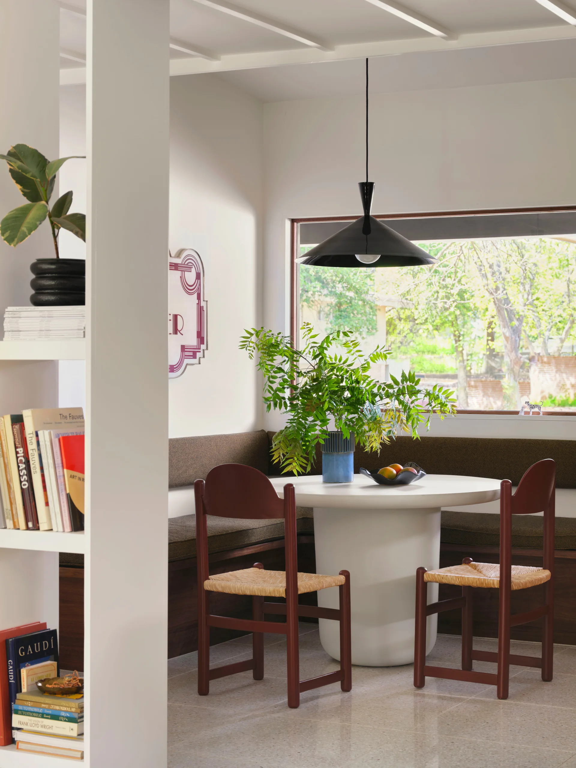 A bright dining nook features a circular white pedestal table, built-in banquette seating with brown cushions, and two dark wood chairs with woven seats.