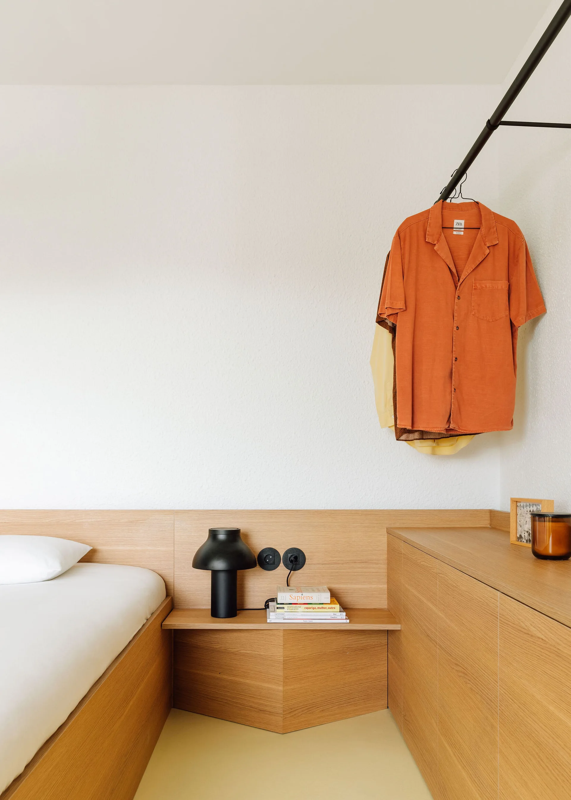 A minimalist bedroom features an integrated wooden bed frame and bedside shelf with a black lamp, next to a built-in dresser and black clothing rack.