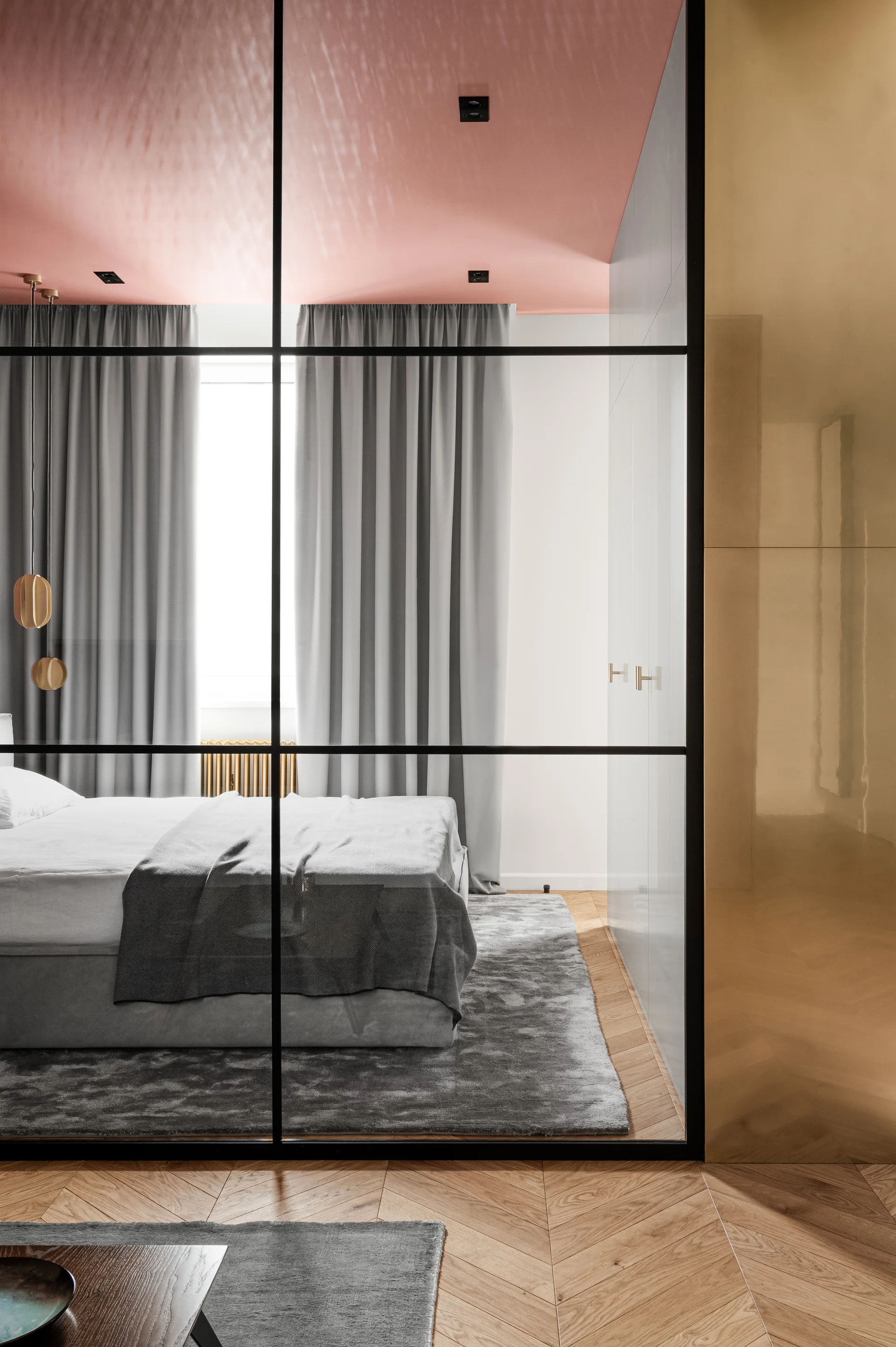 A modern bedroom seen through a black-framed glass partition, featuring a pink painted ceiling, herringbone wood floors, and a bed with gray bedding.