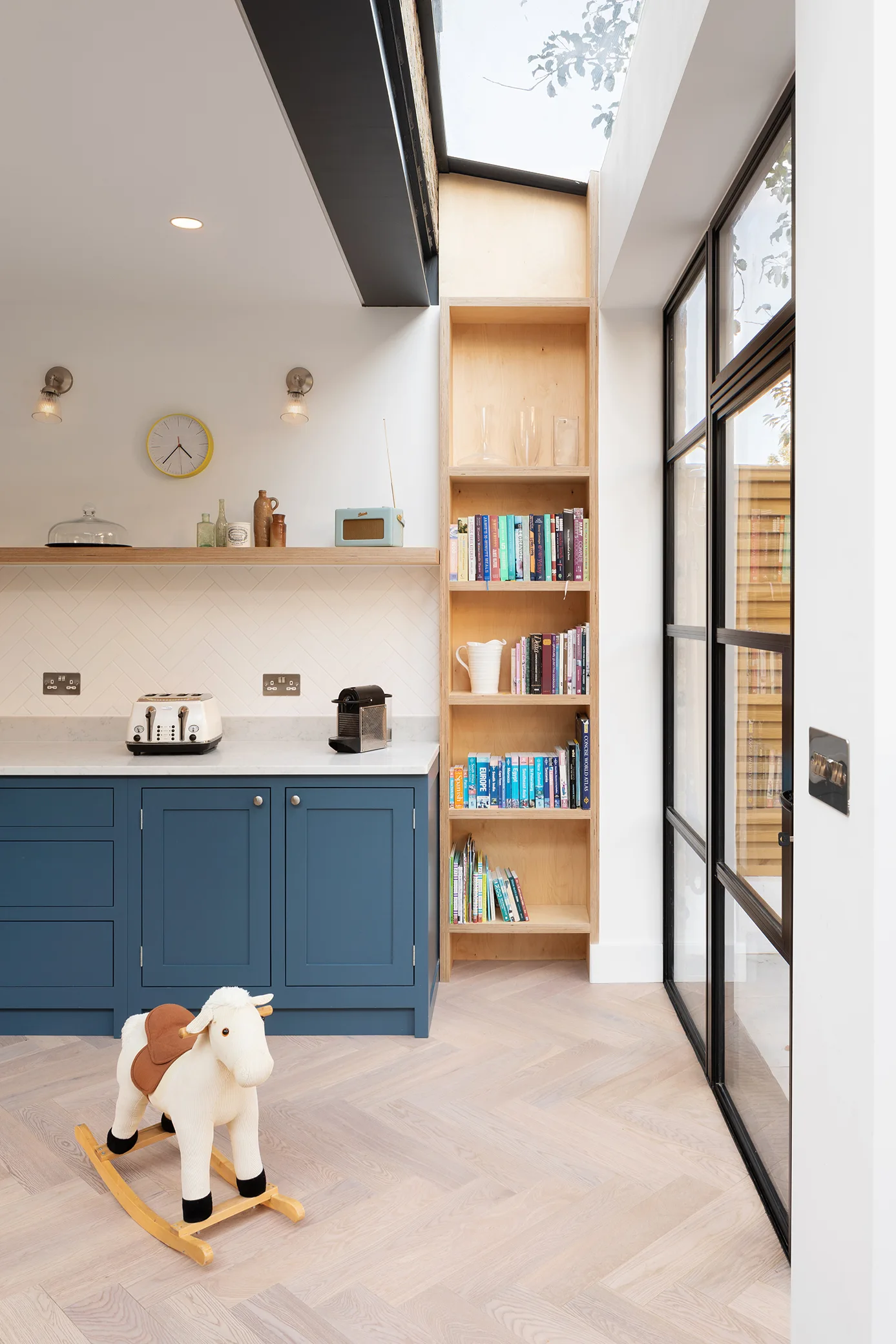 A modern kitchen with blue cabinetry and a tall light wood bookshelf next to a black glass door under a skylight.