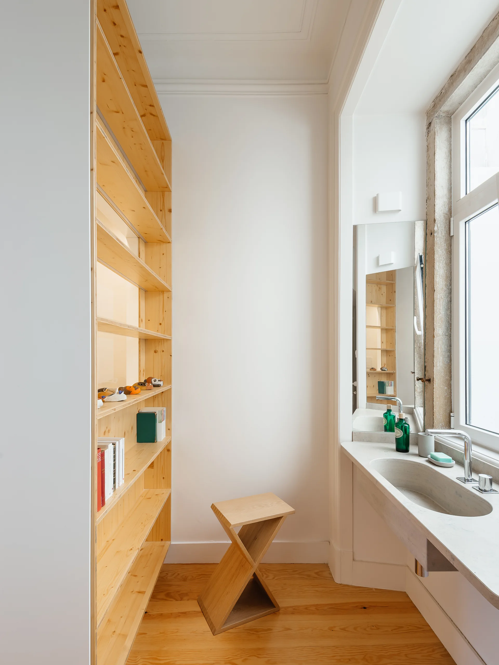 A minimalist bathroom featuring a tall light wood shelving unit, a geometric wooden stool, and an integrated stone-like vanity sink next to a window.