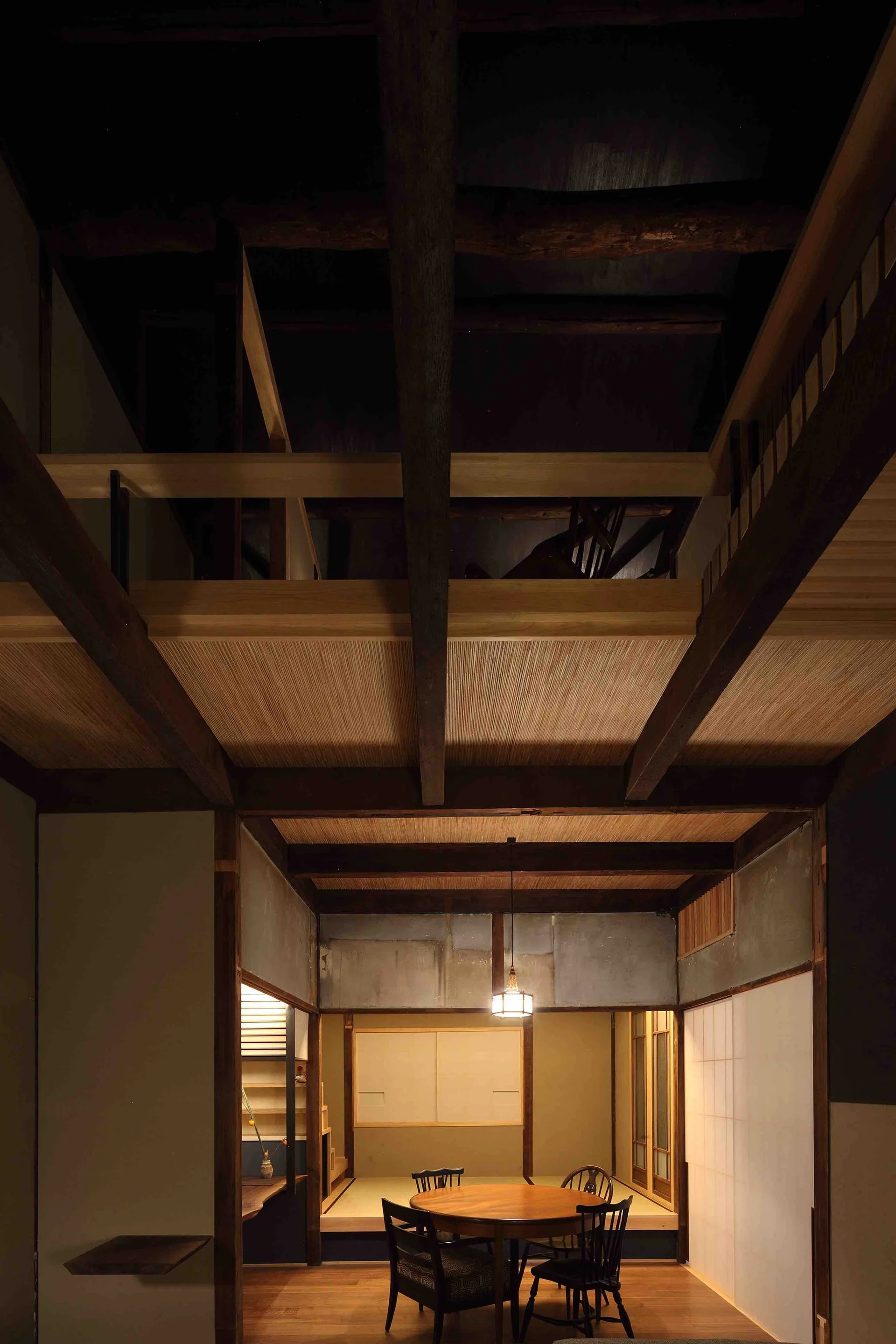 A Japanese dining room with a round wooden table set beneath massive dark timber ceiling beams, featuring tatami mats and a sliding shoji screen behind.