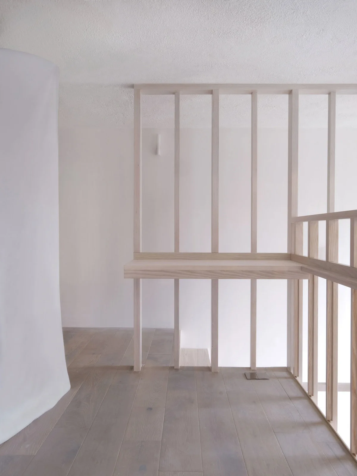 A minimalist staircase landing features a light wood slatted partition, matching wood plank flooring, and smooth white walls.