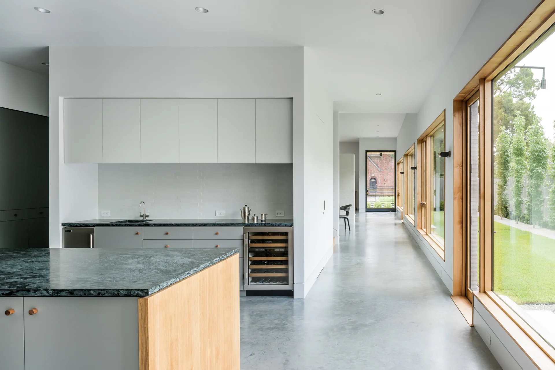 A minimalist kitchen features grey lower cabinets, a dark stone countertop, polished concrete floors, and large wood-framed windows looking out to a garden.