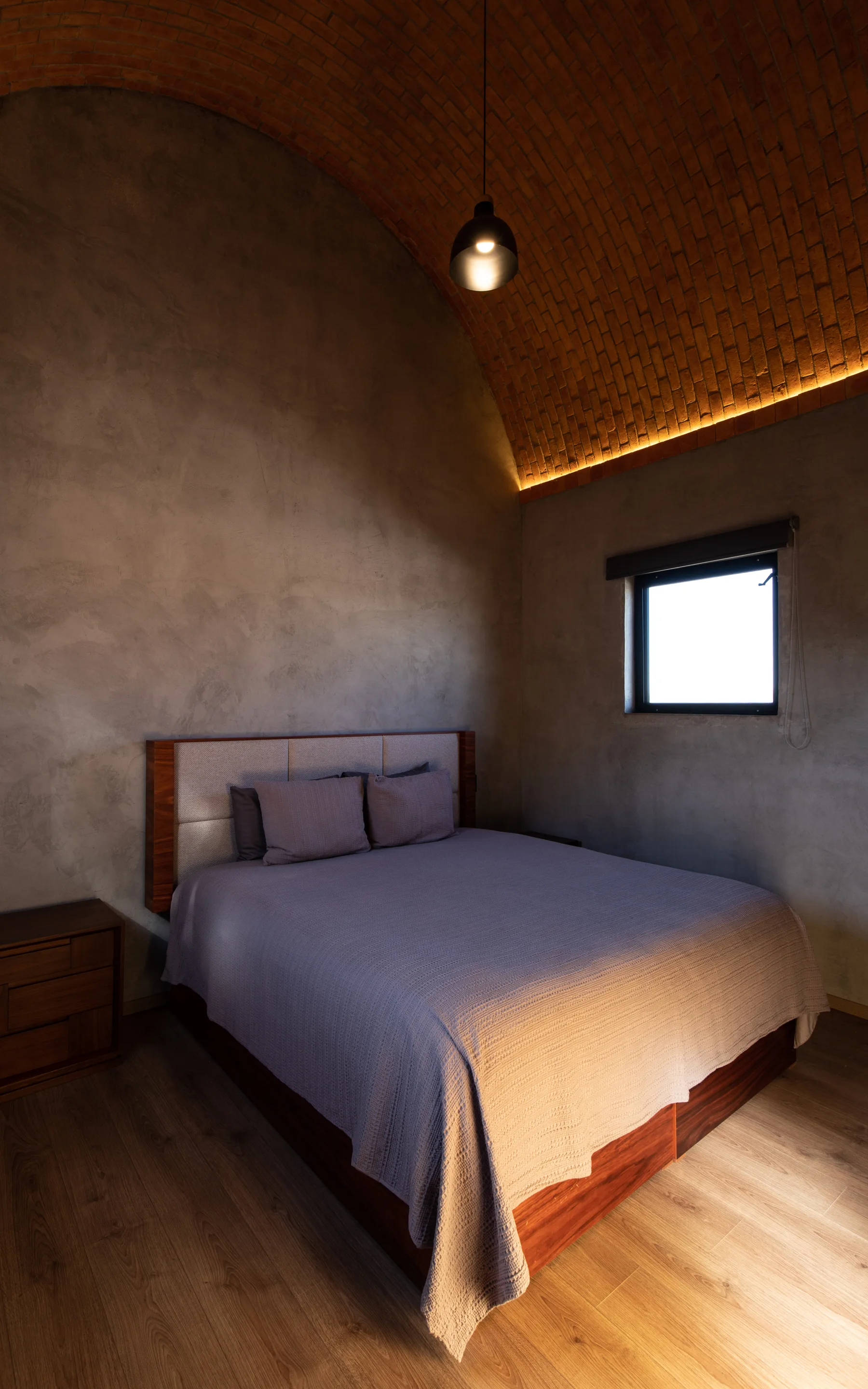 A minimalist bedroom featuring a bed with grey bedding set against textured concrete walls under an arched brick vaulted ceiling.