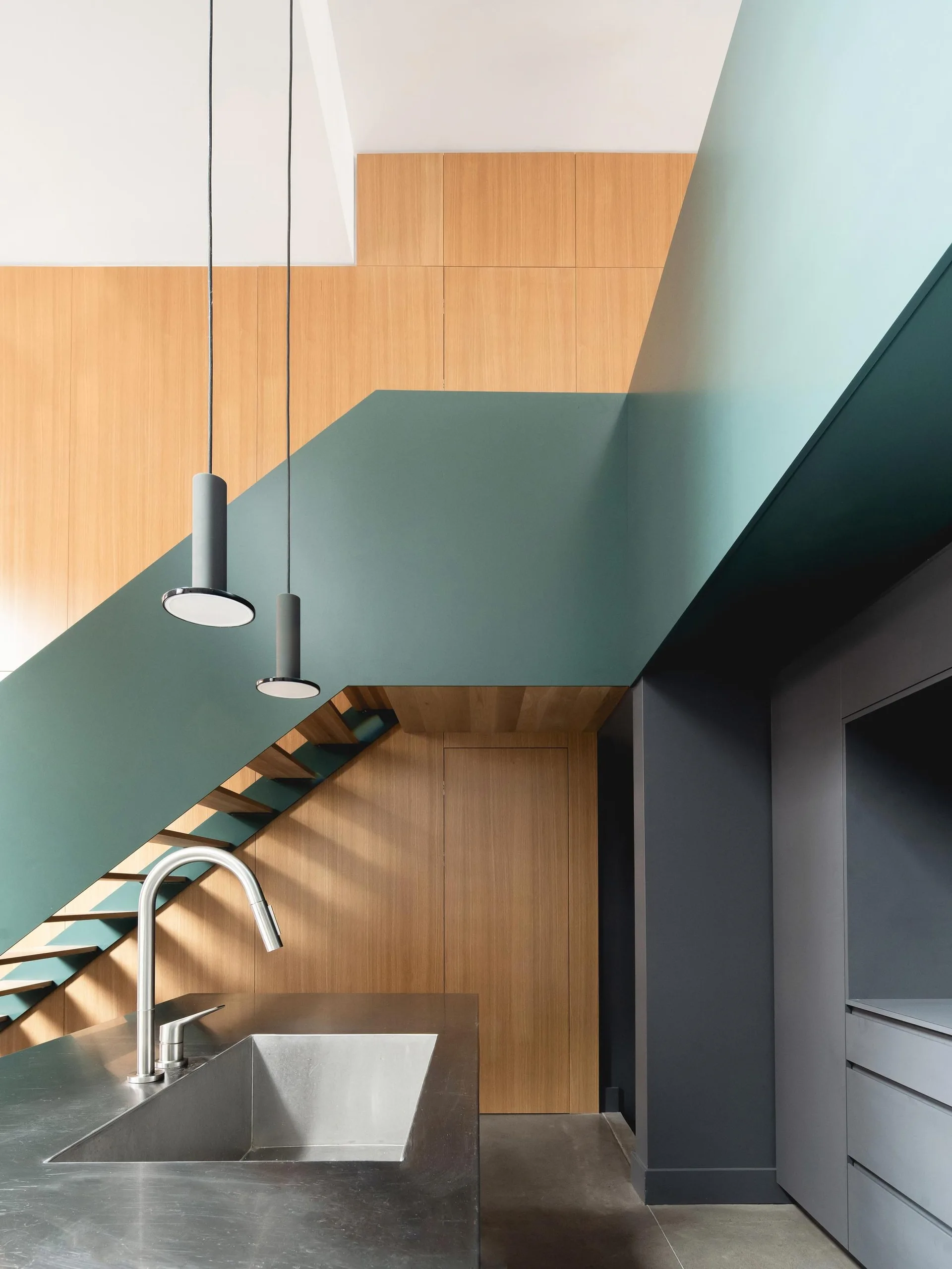 A modern kitchen featuring a dark grey island and sink beneath a prominent teal staircase with light wood wall paneling.