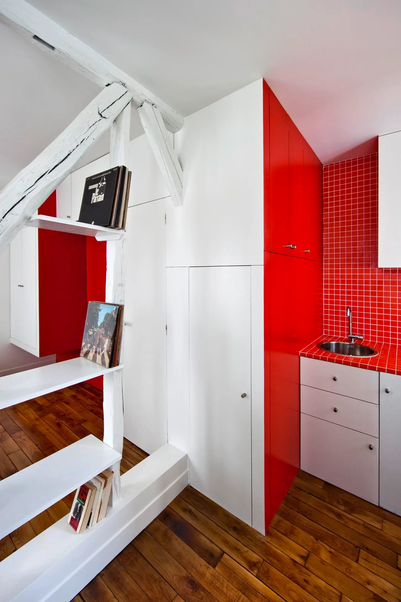 A compact kitchen features glossy red cabinets, a red tiled backsplash, white storage units, and a rustic white timber post with built-in shelves.