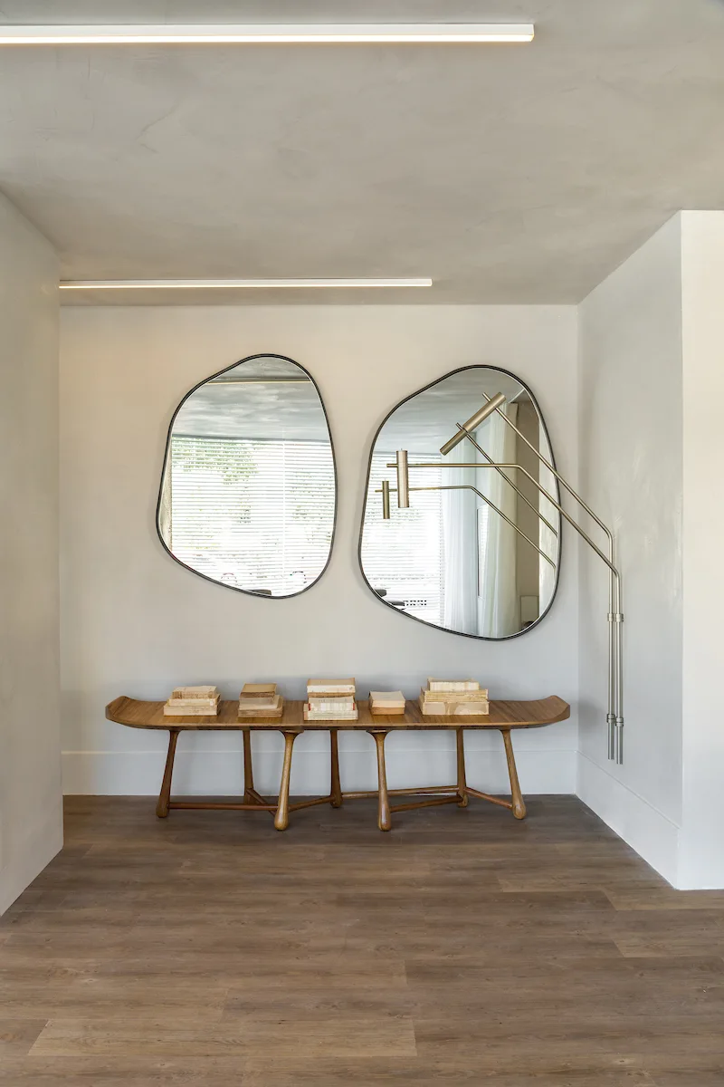 An entryway features a long wooden bench styled with book stacks below two asymmetrical organic-shaped mirrors and a multi-arm brass wall sconce.