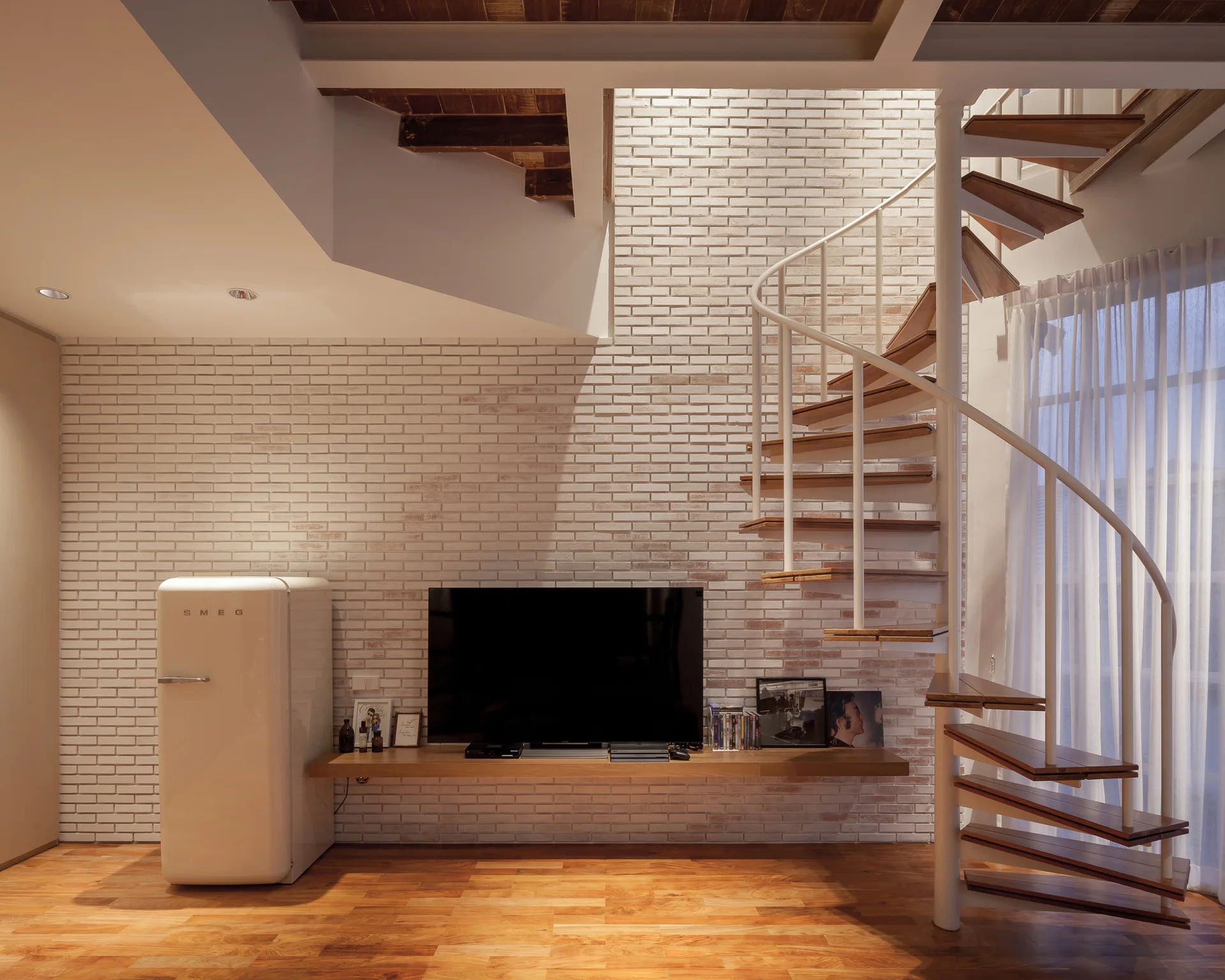 A living room features a white brick feature wall, a white metal spiral staircase, a wood floor, and a retro cream refrigerator next to a TV.