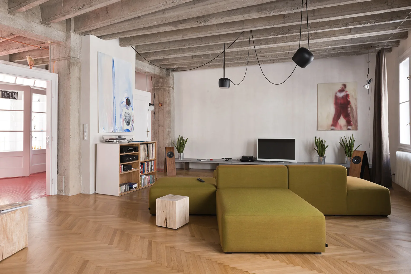 An olive green modular sofa sits on a herringbone wood floor beneath an exposed concrete ceiling in an industrial-style living room.