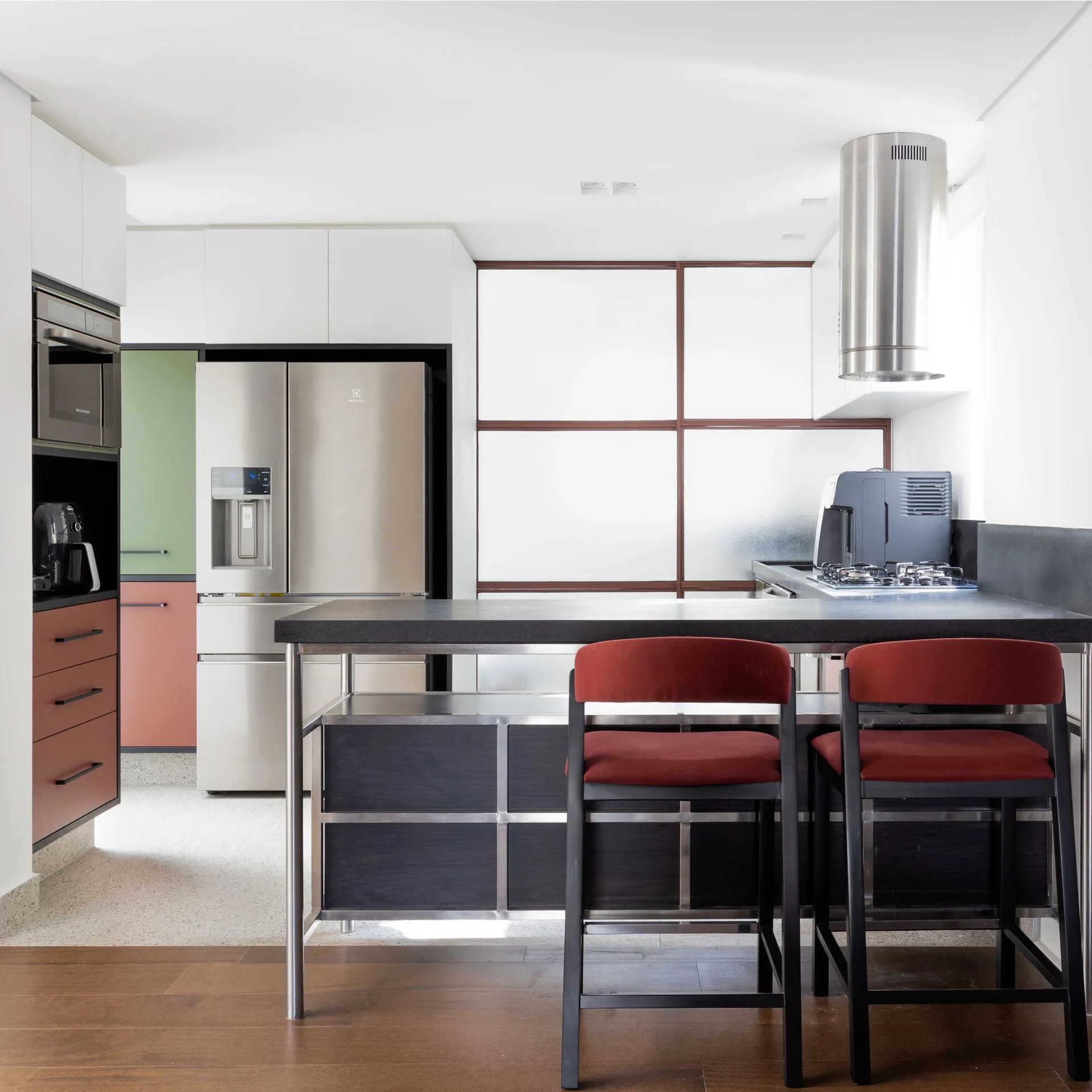 A contemporary kitchen features a dark island with two red stools, stainless steel appliances, and a grid-patterned frosted glass wall.