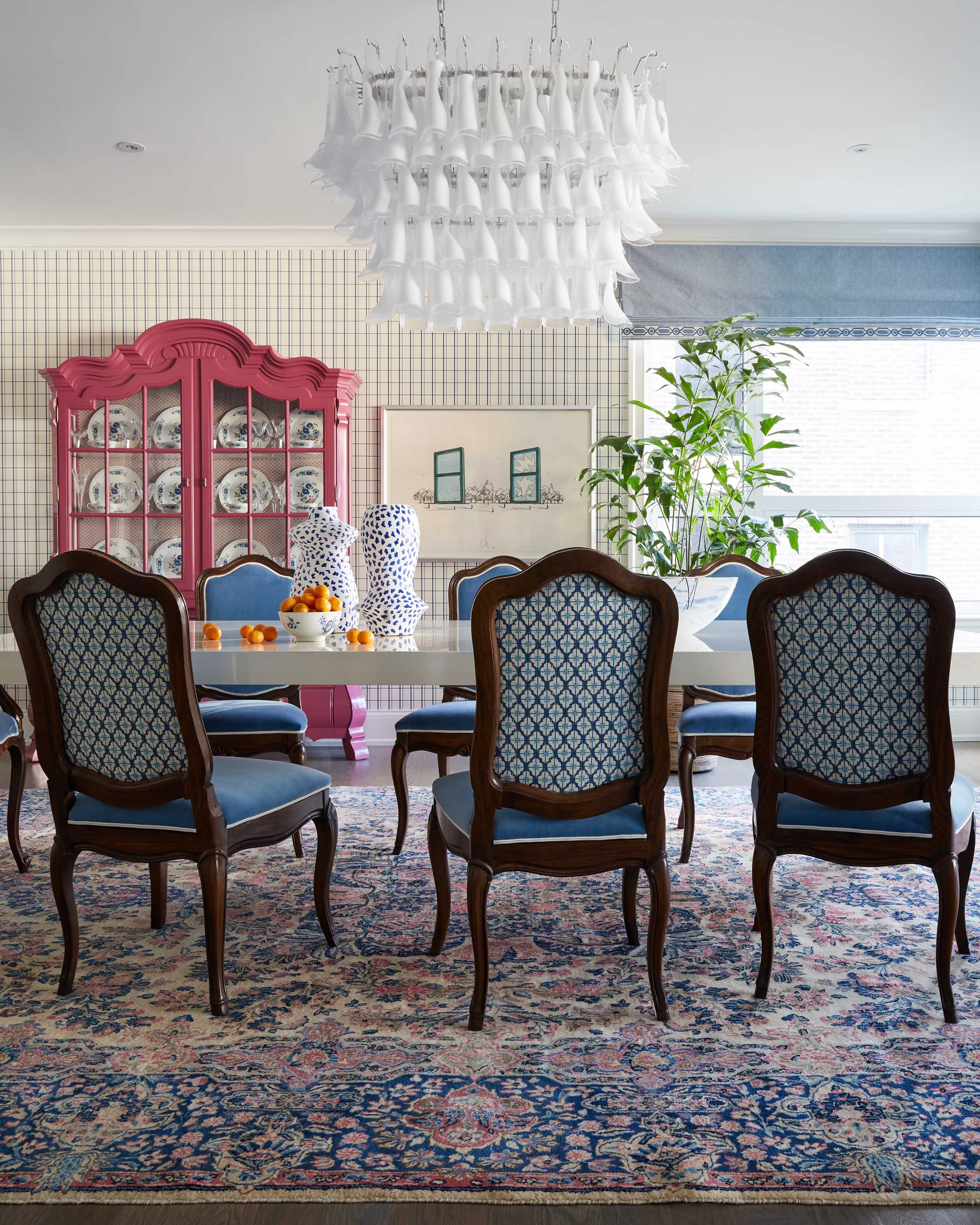 A dining room features a fuchsia china cabinet, blue upholstered chairs around a glossy table, a tiered glass chandelier, and grid pattern wallpaper.