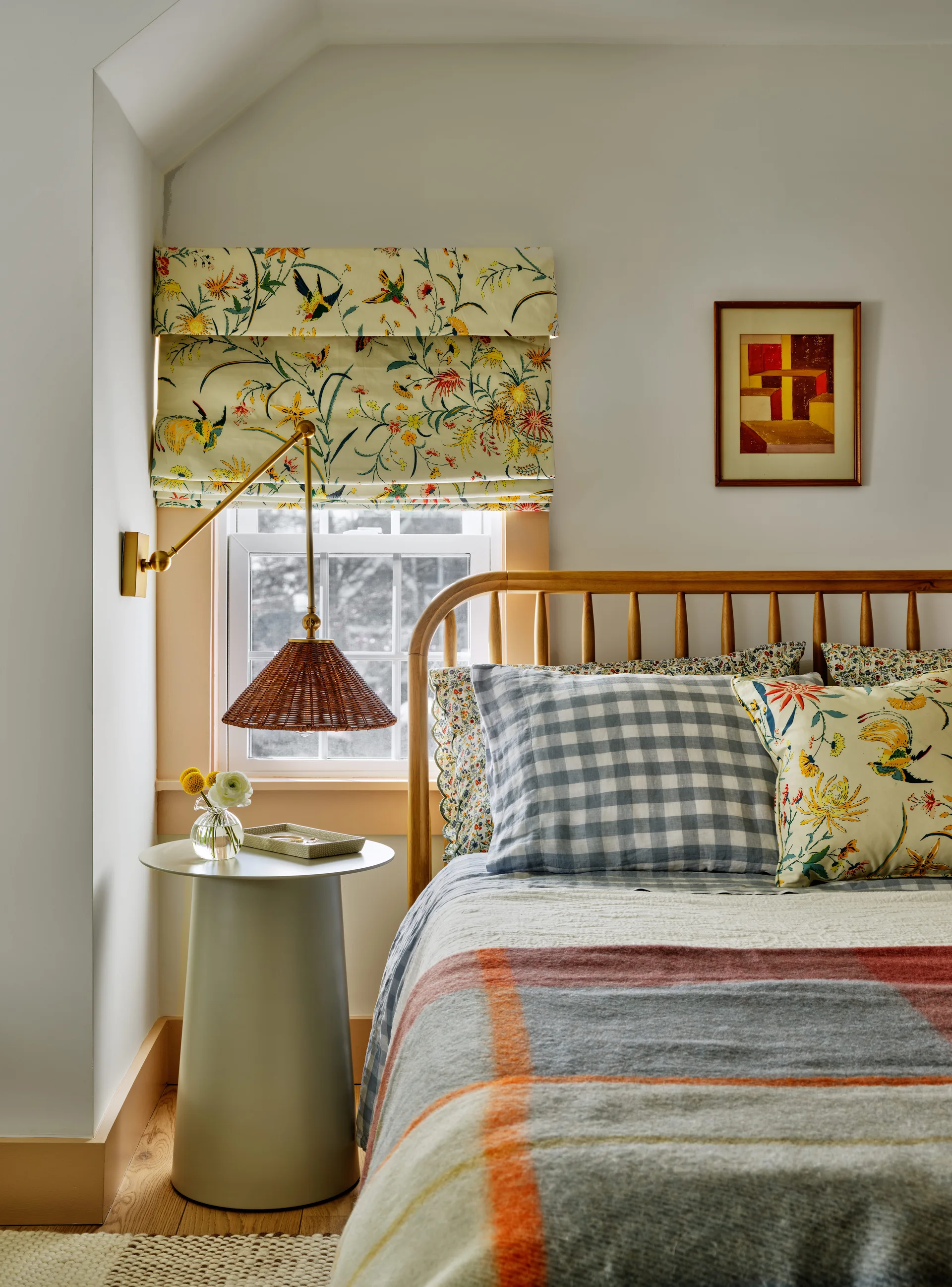 A cozy bedroom corner features a wooden spindle bed with patterned pillows, a metal pedestal nightstand, and a window with a colorful floral roman shade.