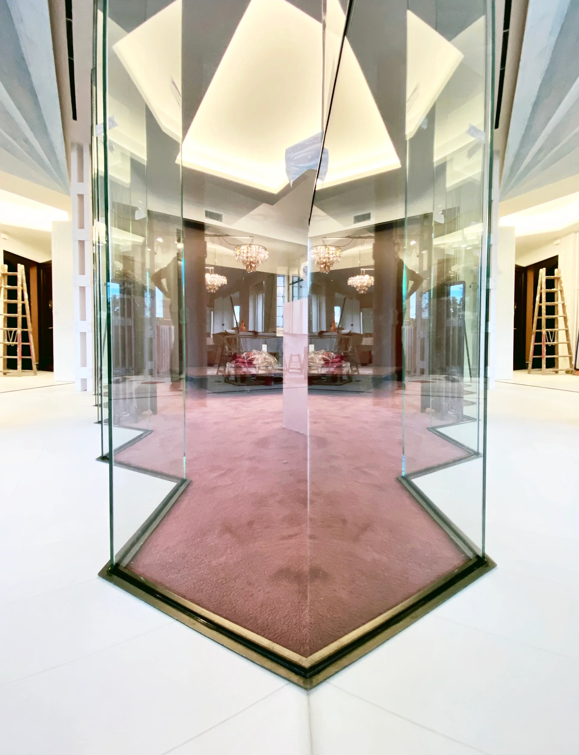 A symmetrical gallery space features an angular glass partition resting on a pink carpet surrounded by white tiled flooring and recessed ceiling lighting.