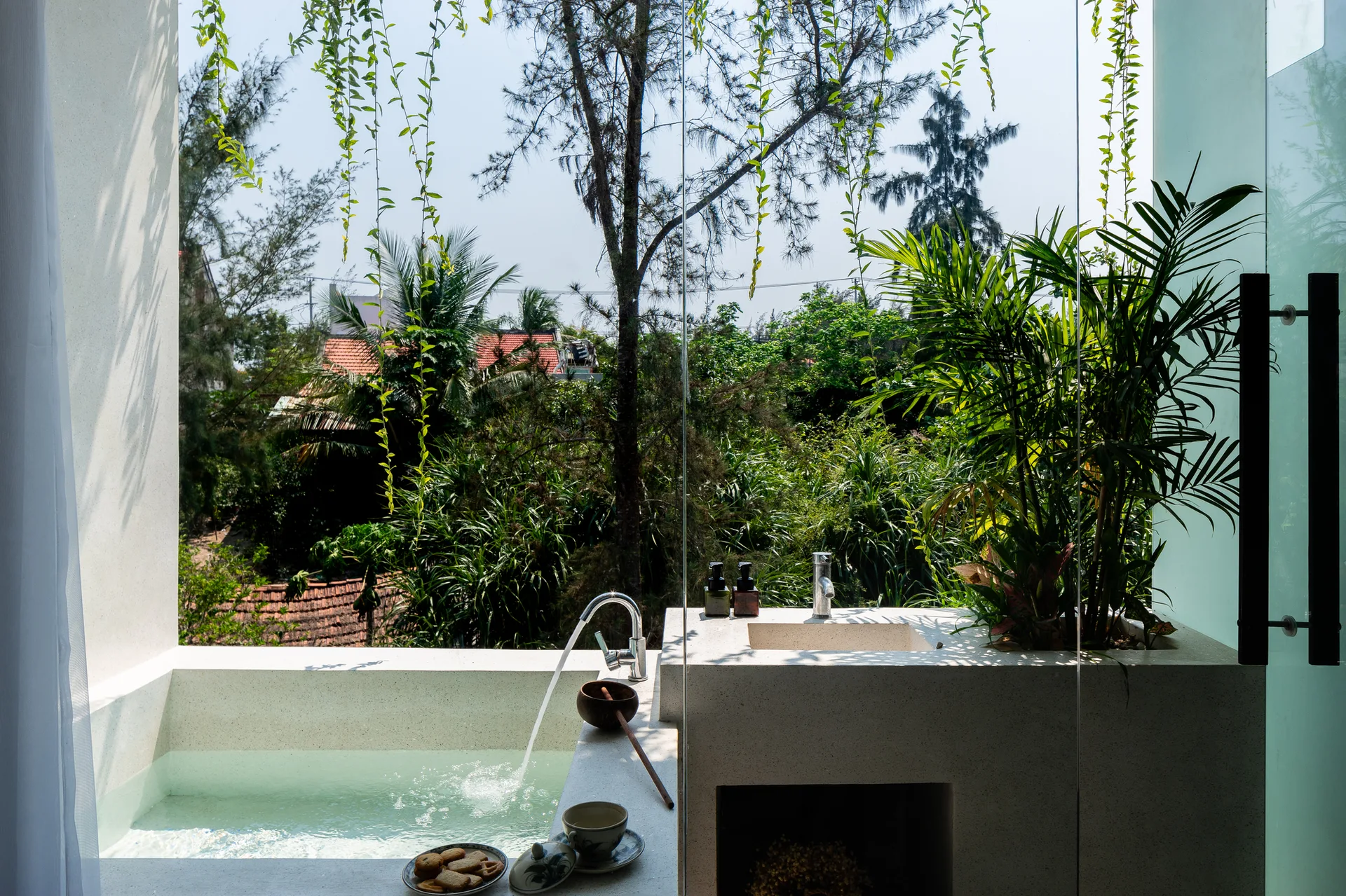 An indoor stone soaking tub and integrated sink vanity sit next to a floor-to-ceiling glass window showing a lush green garden outside.