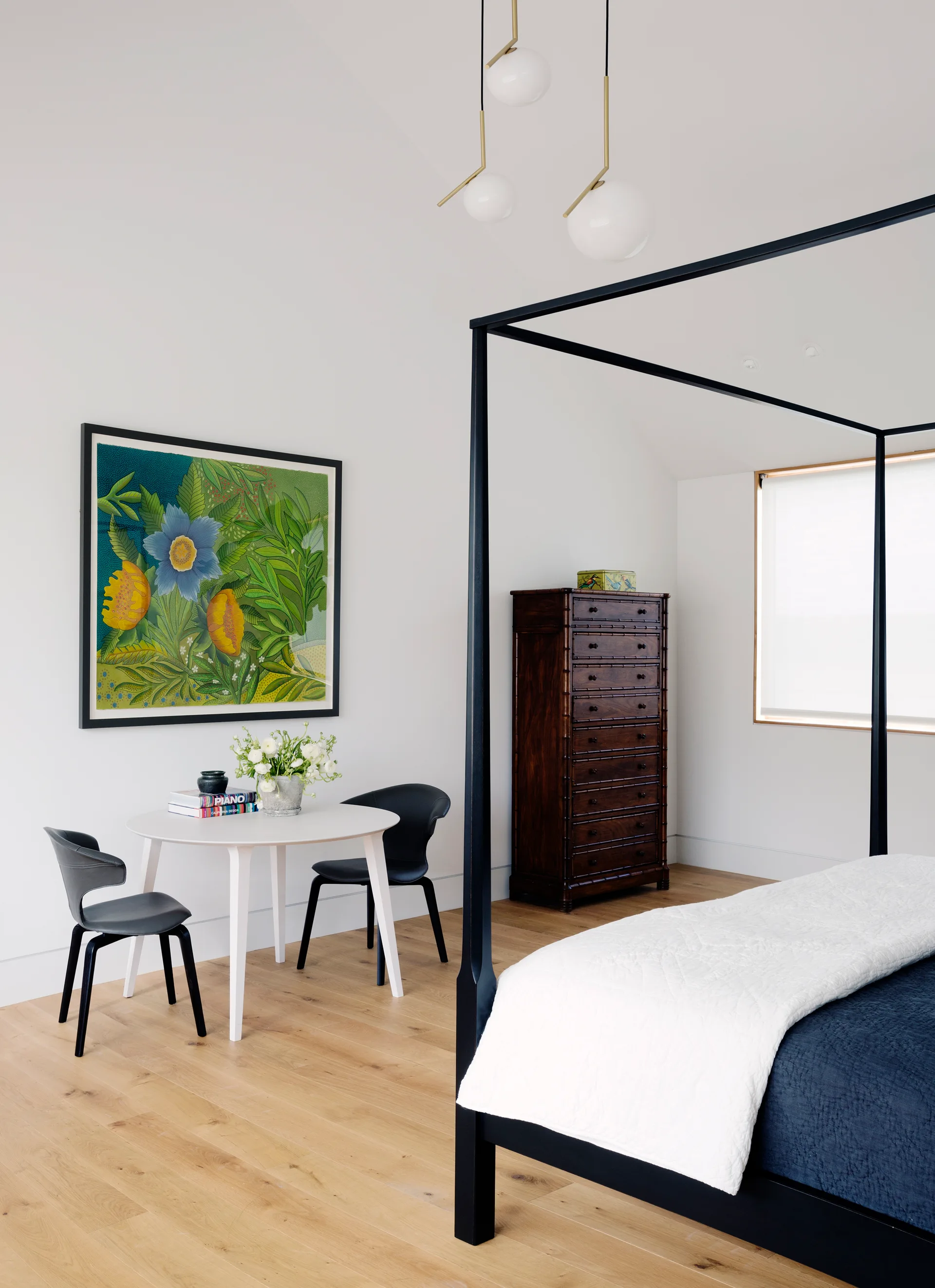 A modern white bedroom features a black canopy bed frame, a small round white table with two black chairs, and a tall dark wood dresser.
